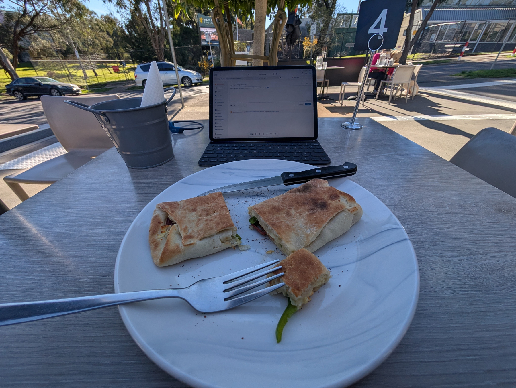 Auto-generated description: A partially eaten sandwich on a white plate with a fork and knife is set on a table outdoors, accompanied by a tablet displaying text.