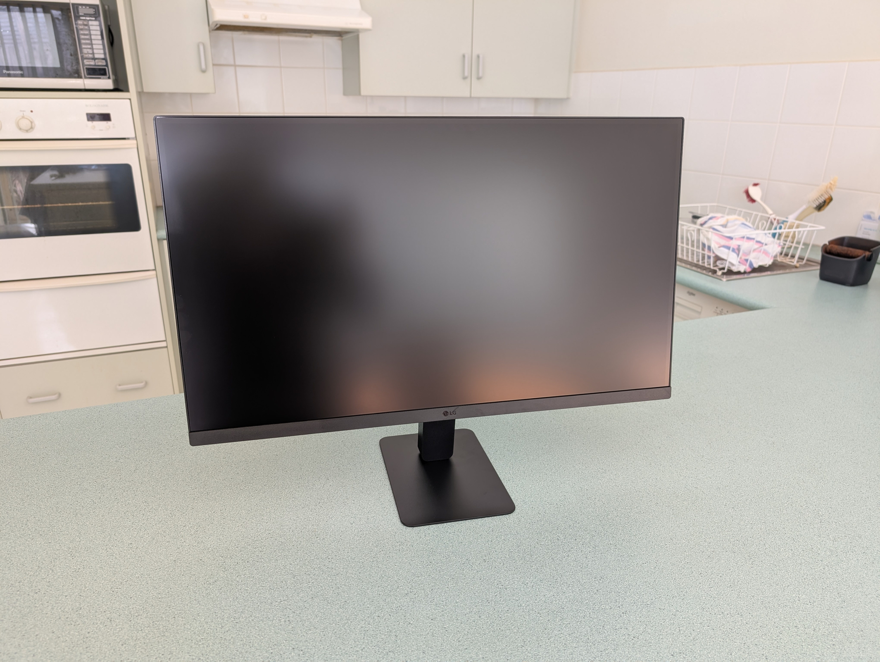 Auto-generated description: A widescreen computer monitor is placed on a kitchen bench with cabinets, an oven, and kitchen items visible in the background.