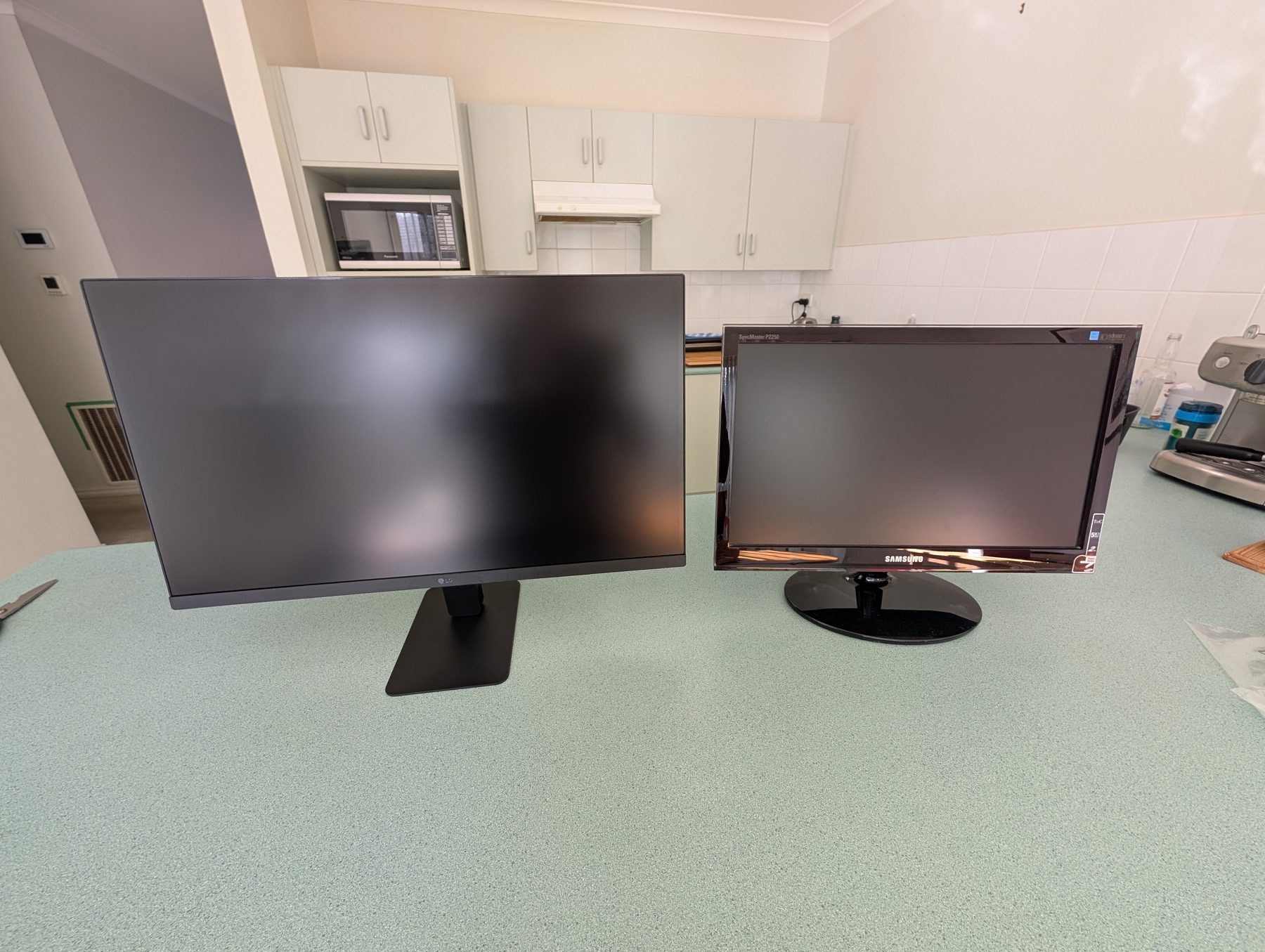 Auto-generated description: Two computer monitors of different sizes are placed side by side on a bench in a kitchen setting.
