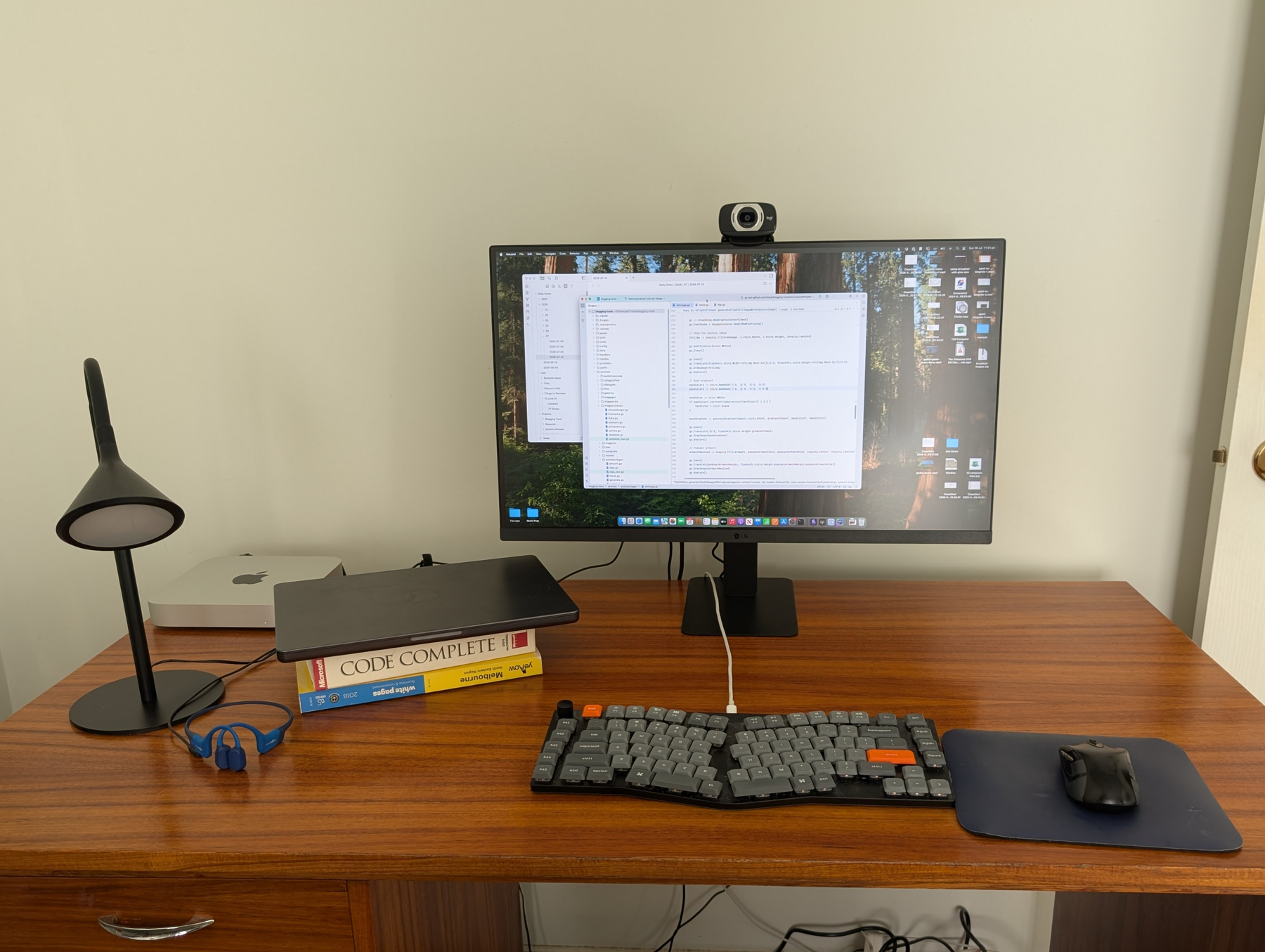 Auto-generated description: A computer monitor displaying coding software is placed on a wooden desk with an ergonomic keyboard, mouse on a mousepad, a desk lamp, a pair of blue glasses, and a stack of books including Code Complete.