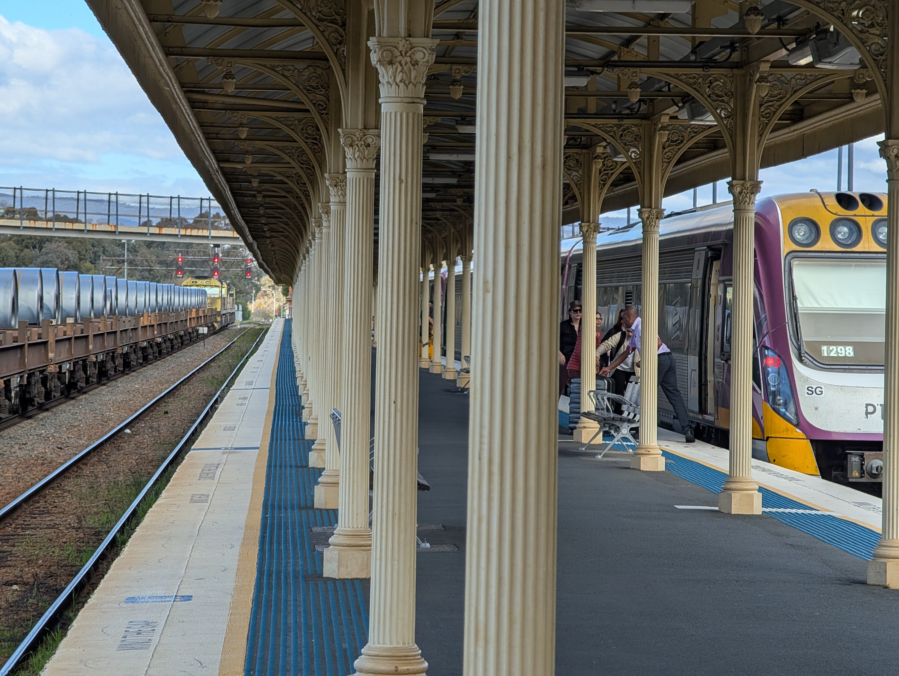 Auto-generated description: A train is stopped at a platform with ornate columns under a covered waiting area while another train is passing by on adjacent tracks.