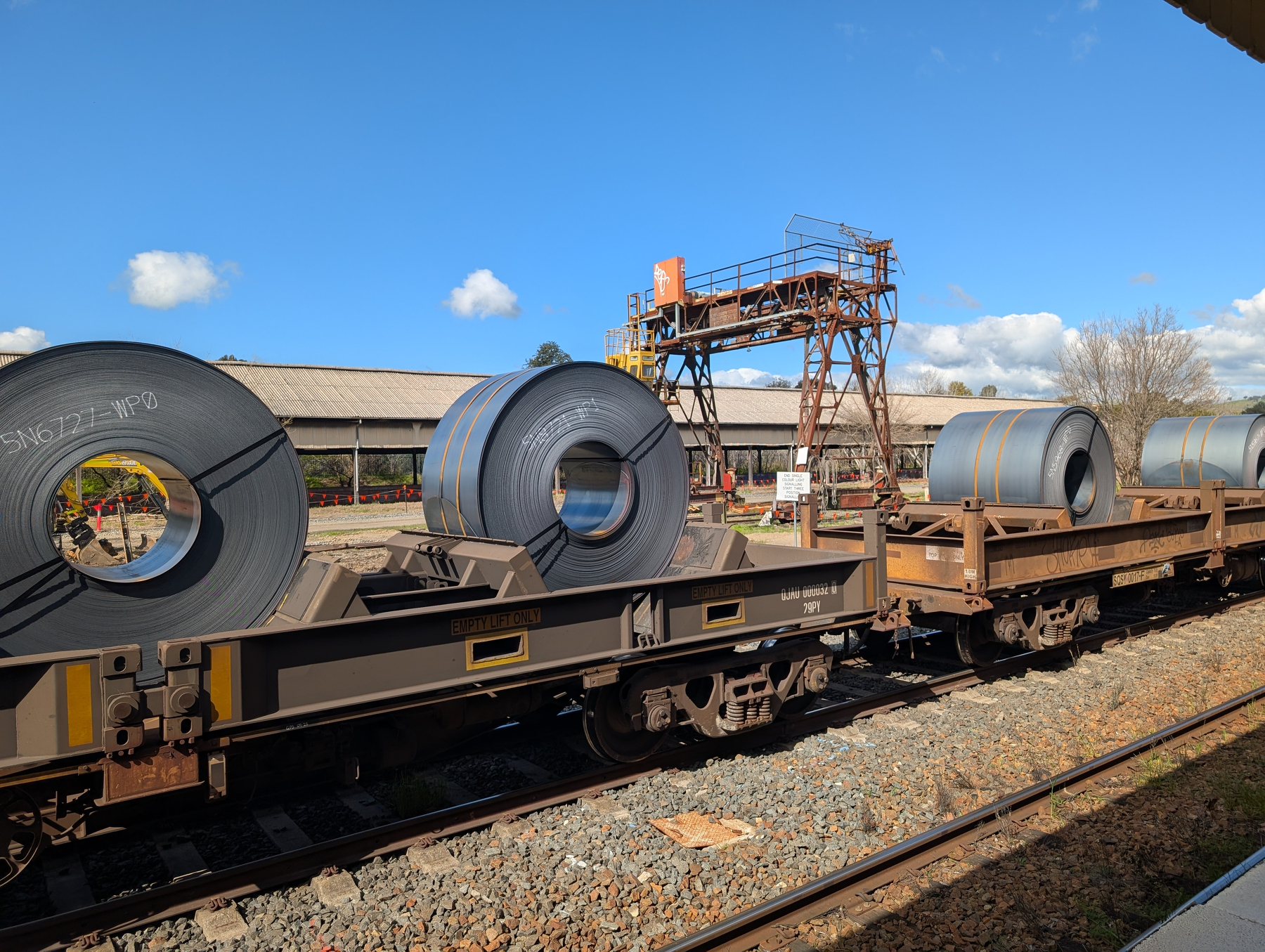 Auto-generated description: Large rolls of steel are being transported on an open flatbed train under a clear blue sky.