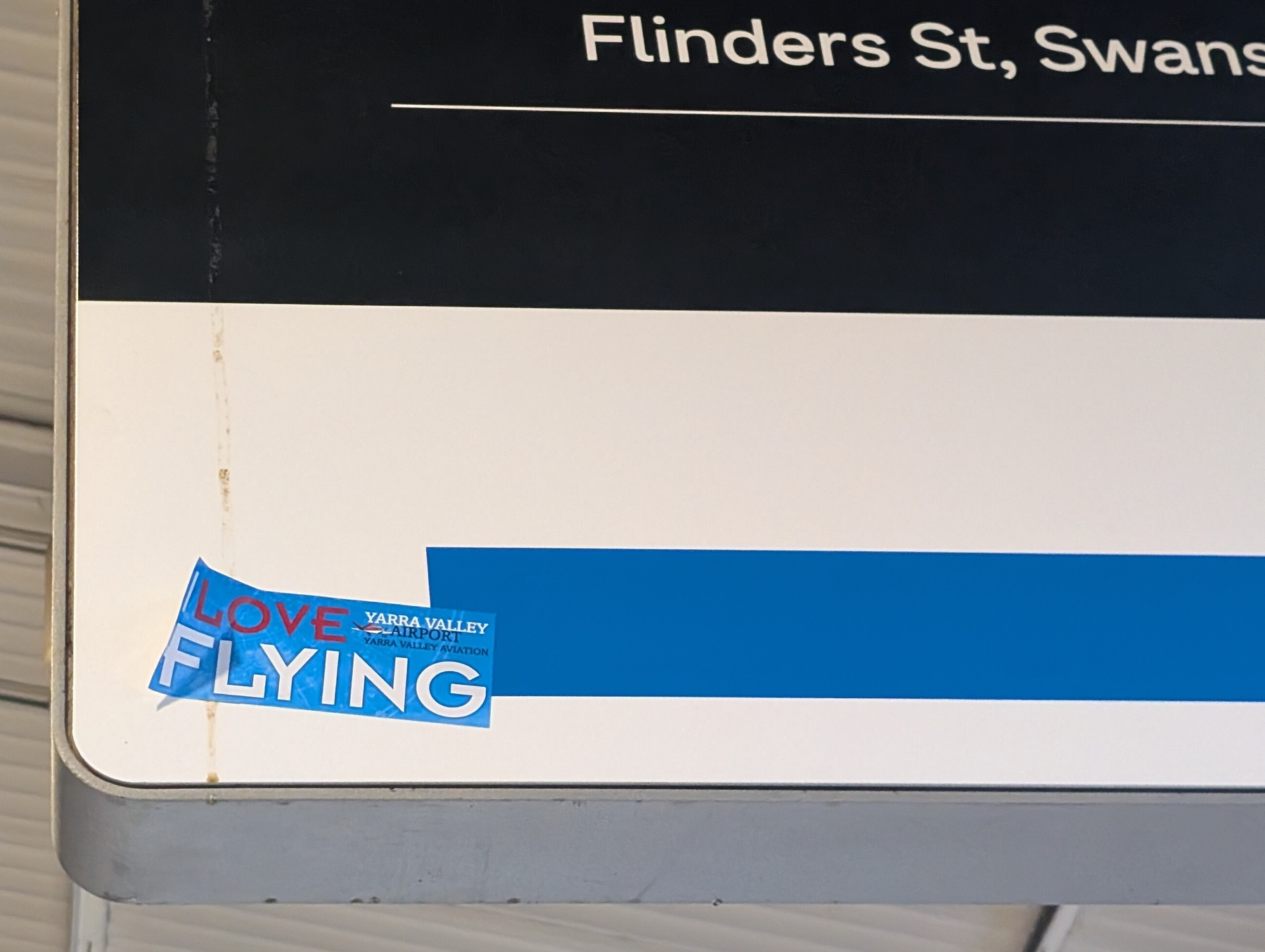 A partial street sign displays the text Flinders St, Swans with a small blue sticker at the bottom left corner saying LOVE FLYING and referencing Yarra Valley Transport.