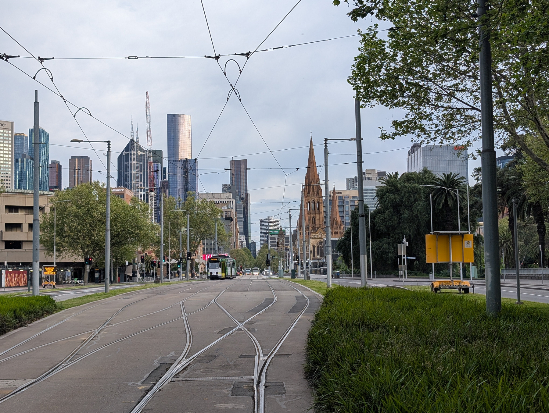 Auto-generated description: A city street with tram tracks leads toward a skyline of modern skyscrapers and a historic church with a pointed steeple.