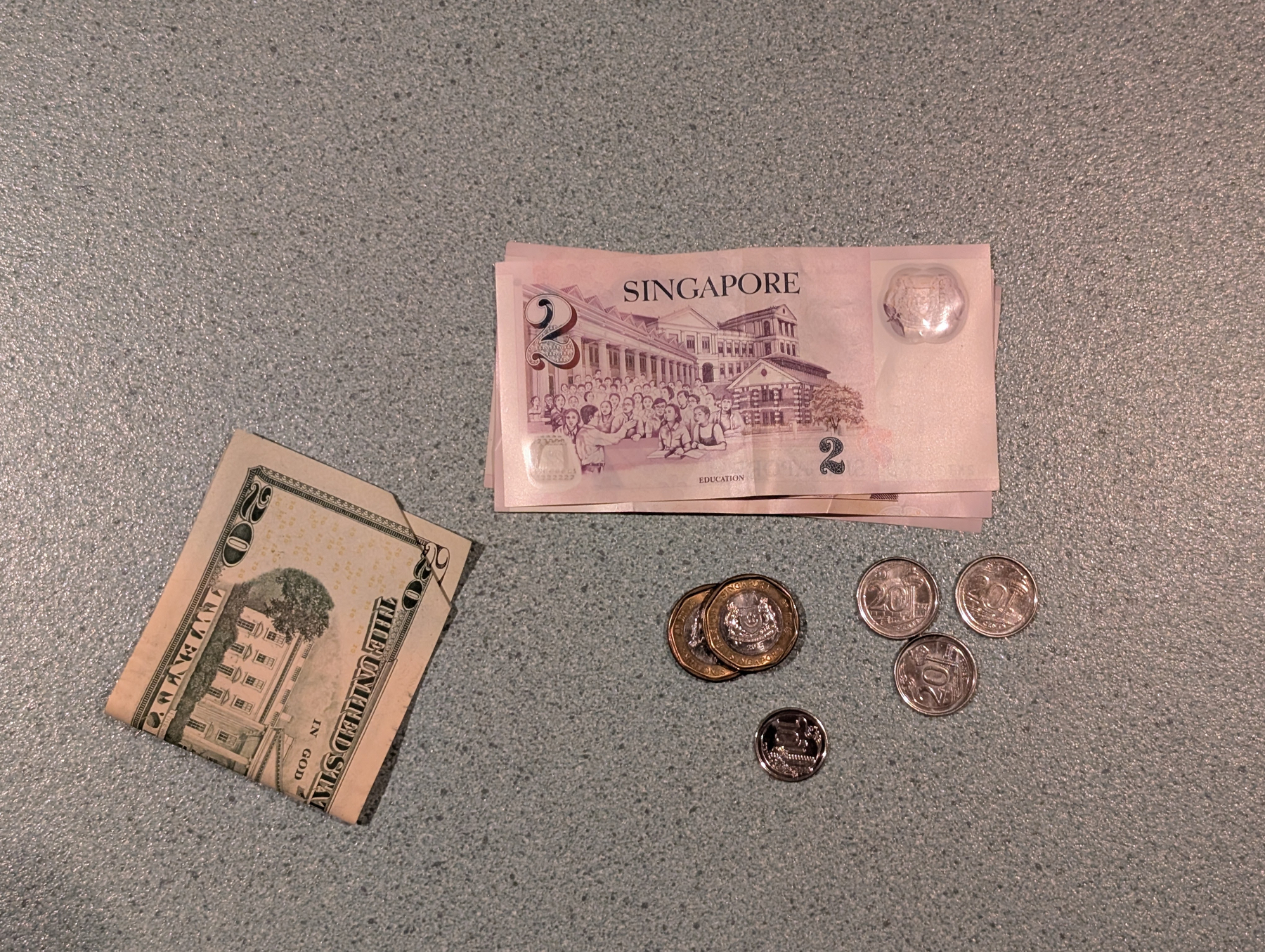 Auto-generated description: A collection of Singapore currency including pink two-dollar bills and various coins is displayed on a gray surface alongside a folded twenty-dollar bill.