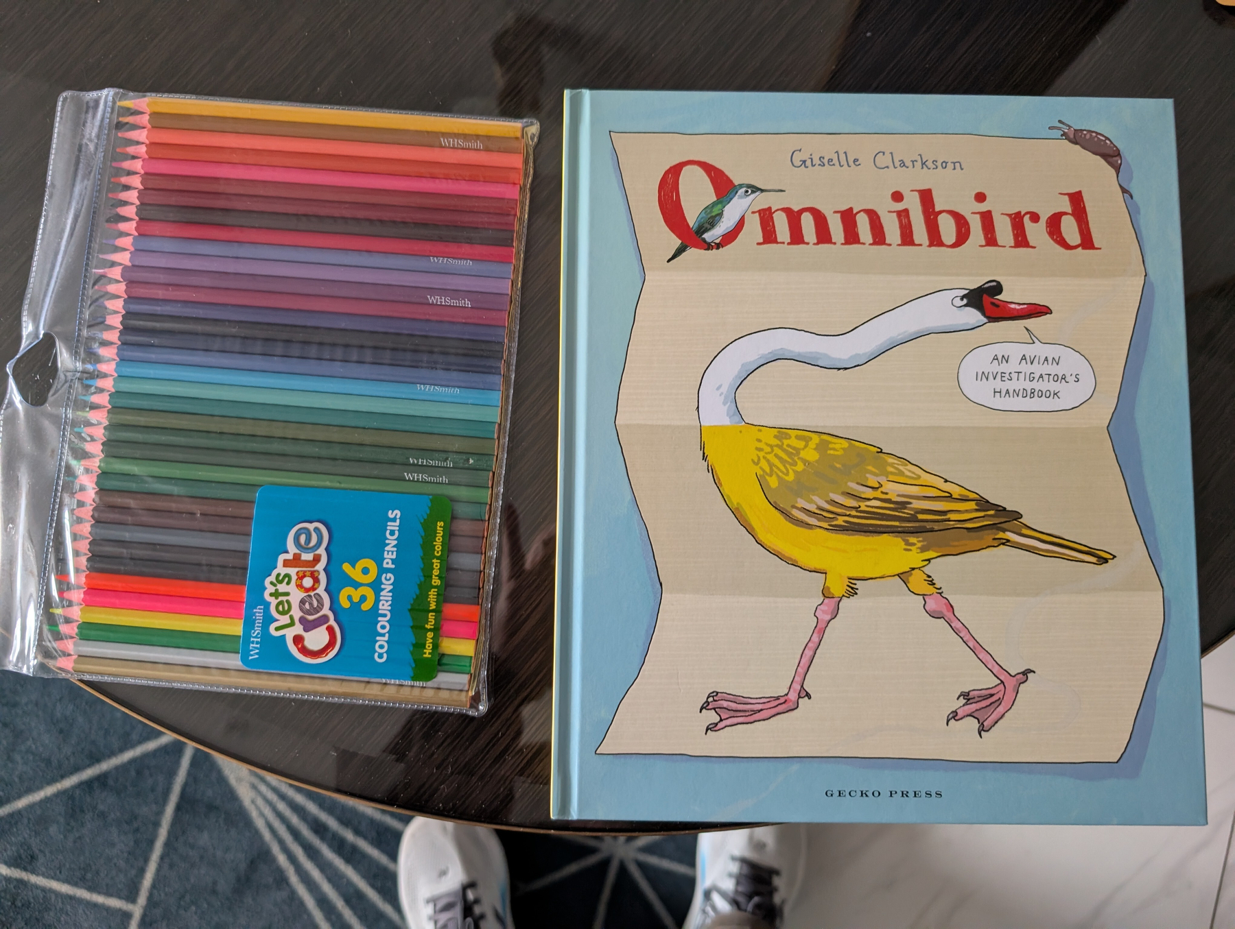 Auto-generated description: A set of 36 colorful pencils and a book titled Omnibird: An Avian Investigator's Handbook by Giselle Clarkson are placed on a dark round table.