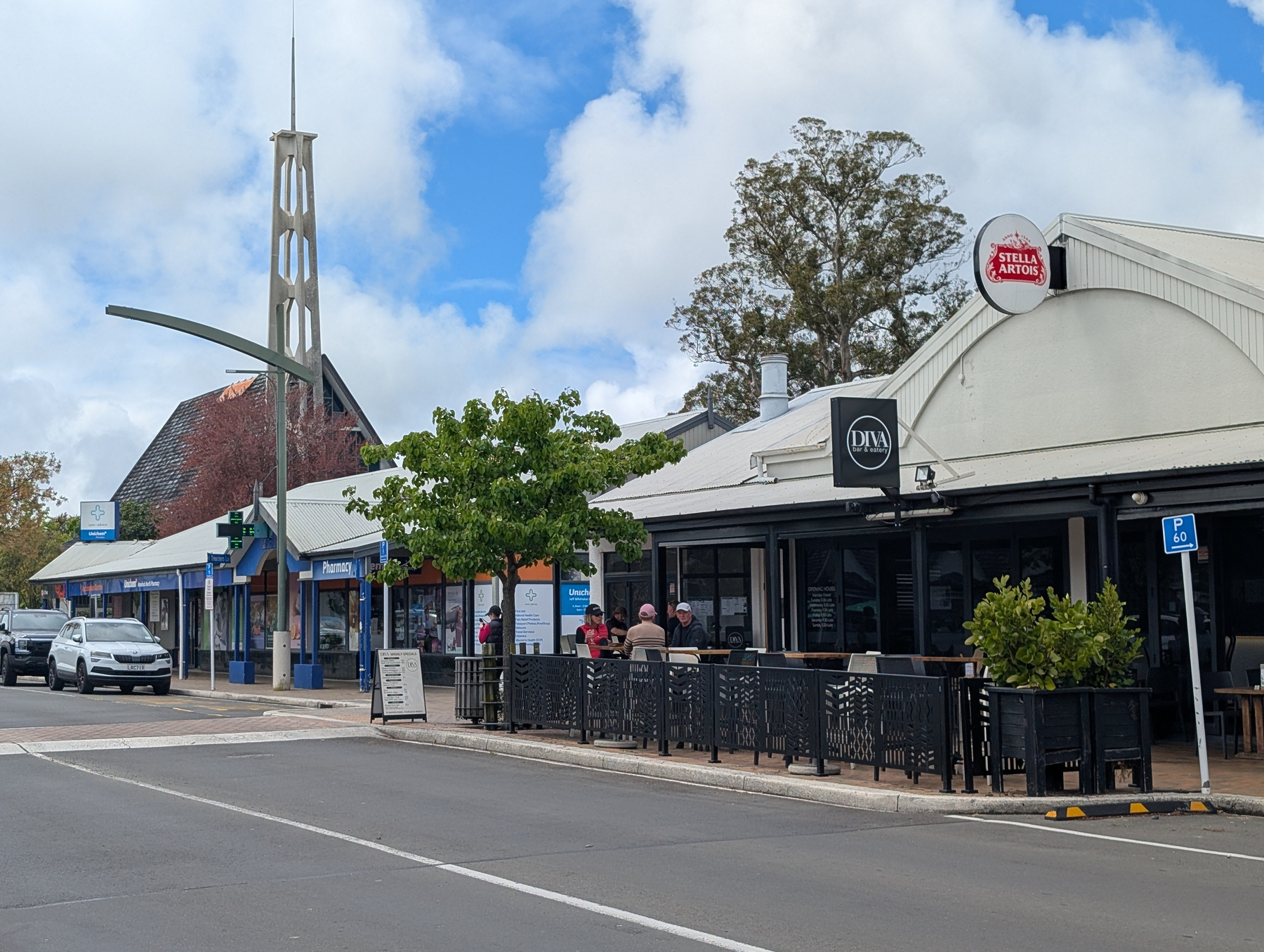 Auto-generated description: A street scene shows a row of shops and restaurants with outdoor seating and a distinctive tall tower structure in the background under a partly cloudy sky.