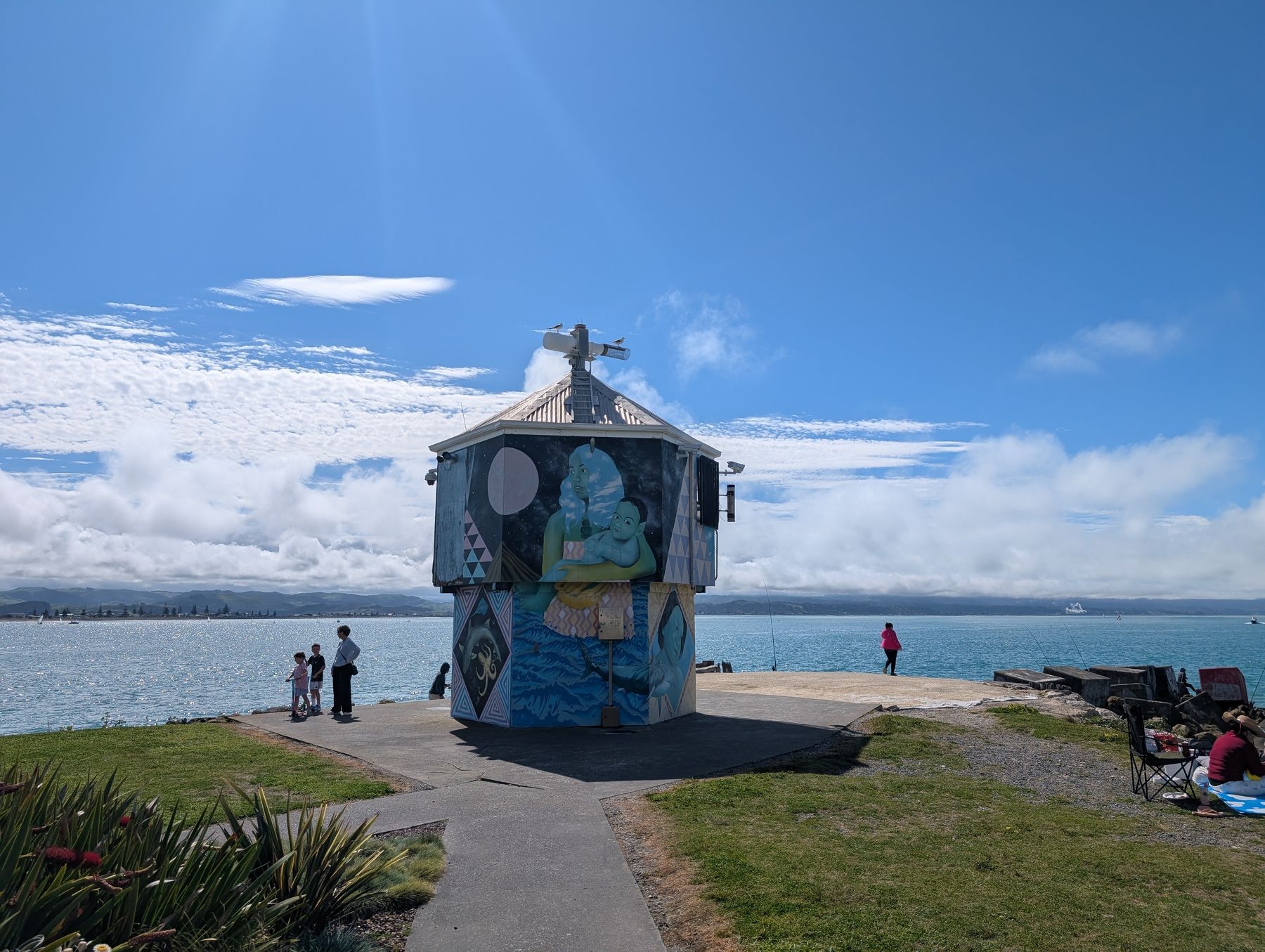Auto-generated description: A small, painted pavilion stands on a waterfront promenade where people are enjoying the sunny day near the calm sea.
