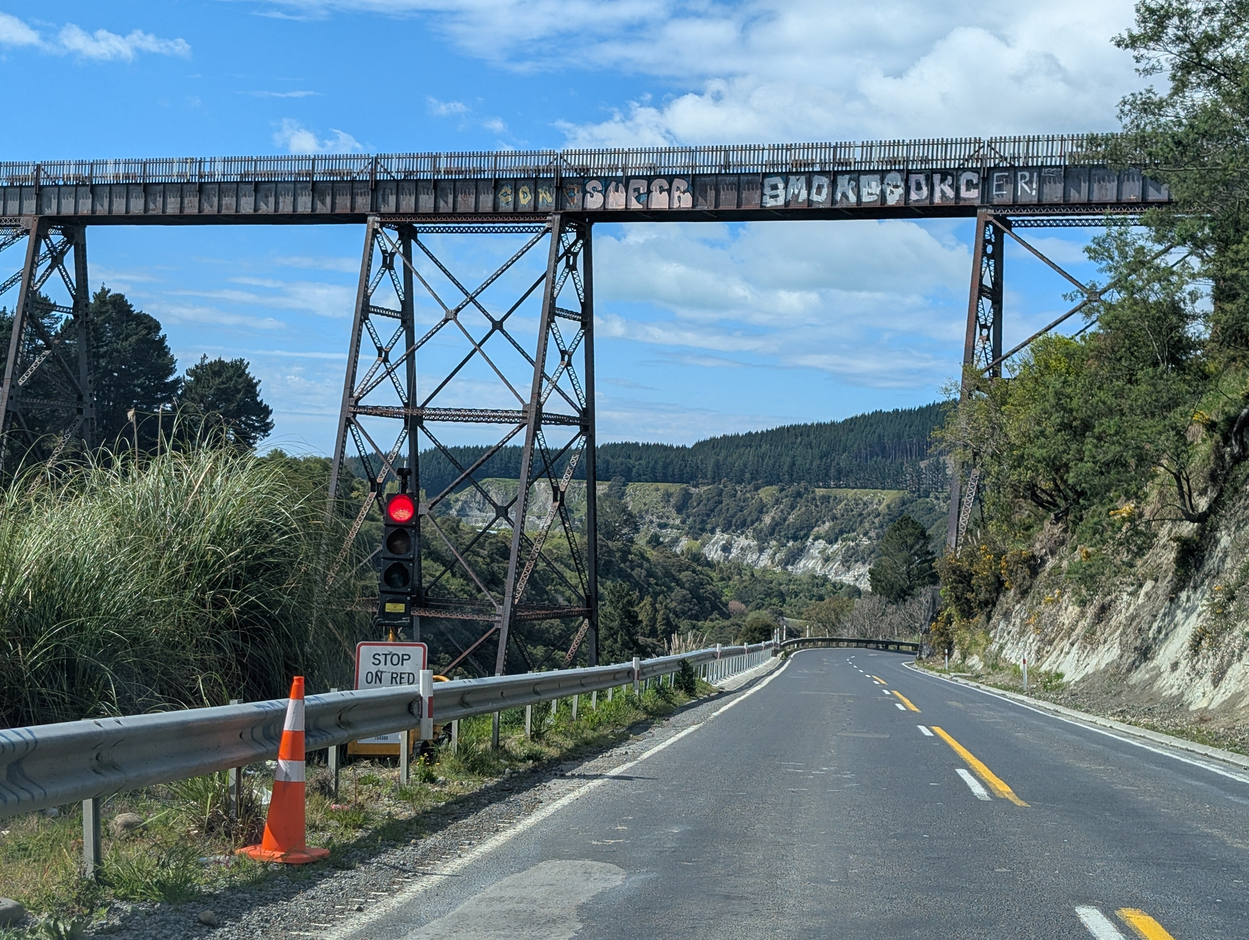 Auto-generated description: A road passes under a tall railway bridge with graffiti, while a red traffic light signals to stop, and a sign below reads STOP ON RED.