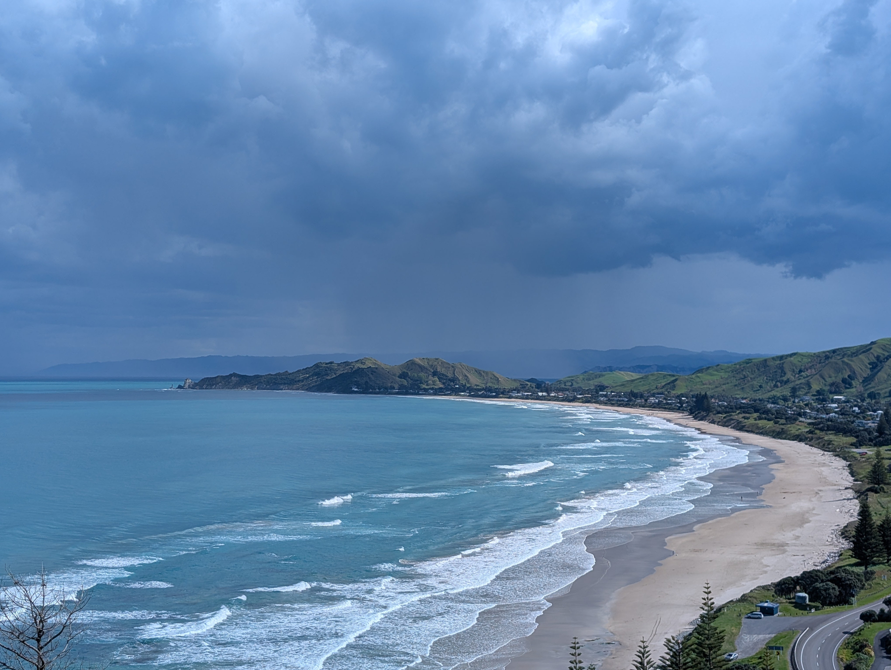 Auto-generated description: A coastline with gentle waves rolling onto a sandy beach is bordered by green hills under a cloudy sky.
