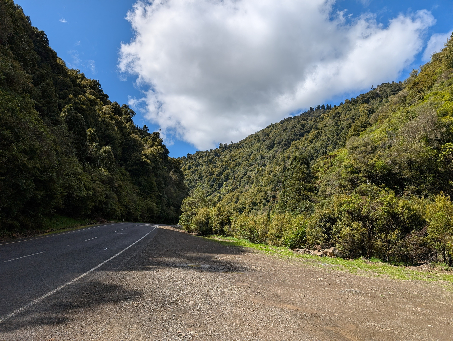 Auto-generated description: A winding road runs alongside green, forested hills under a partly cloudy blue sky.