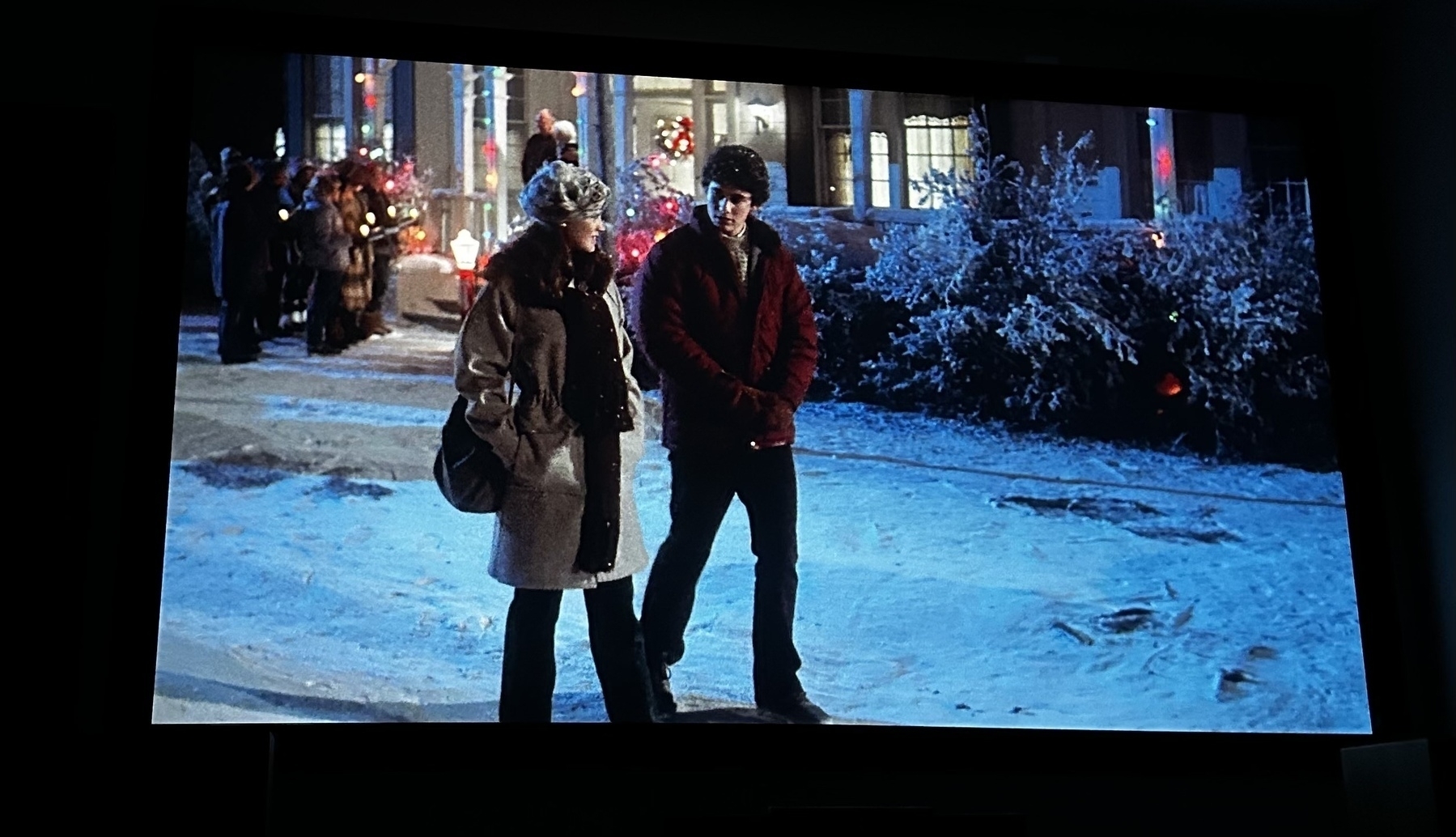 Two characters walking through a Christmas scene with carolers in the background. 