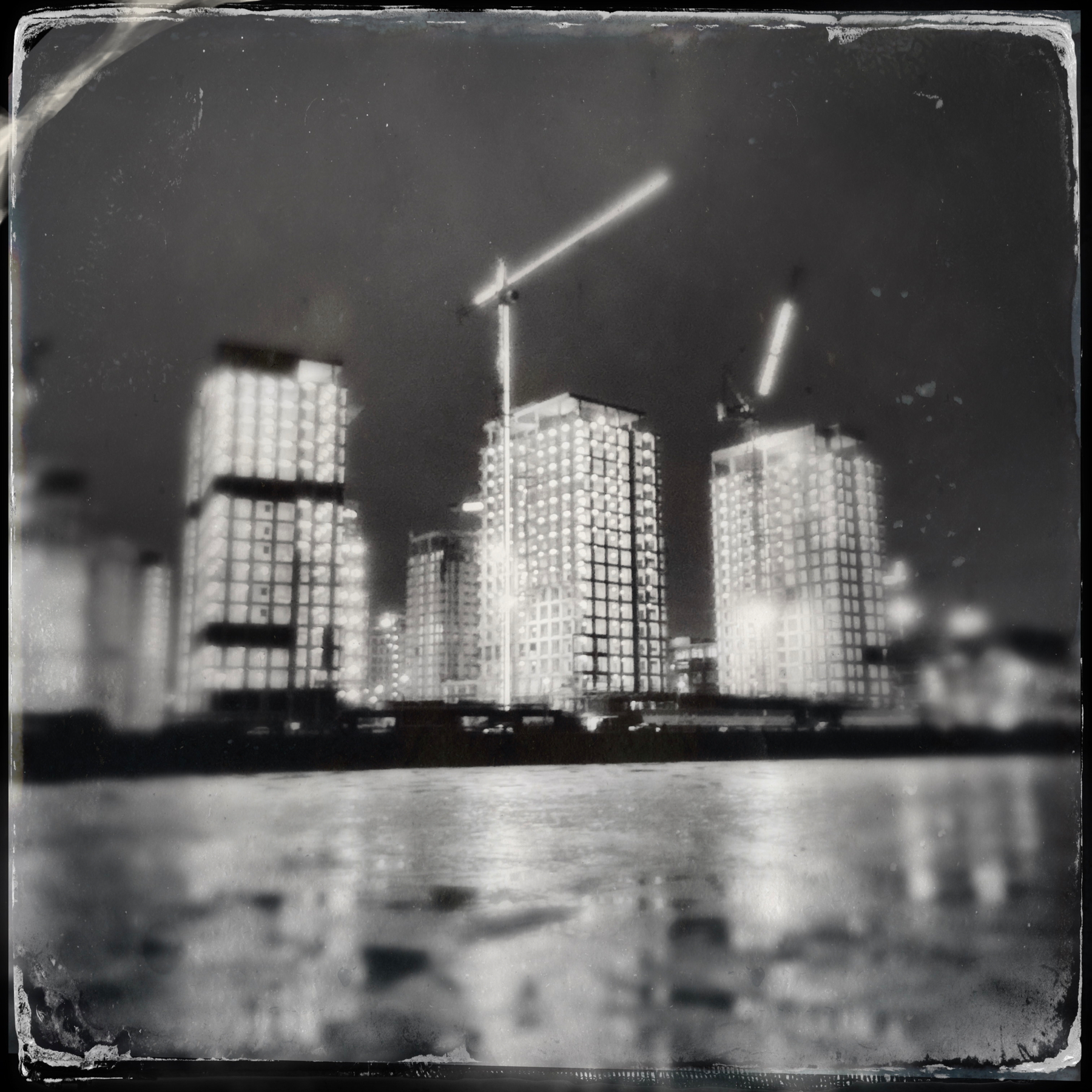A nighttime cityscape showcases tall buildings under construction with cranes, reflected in a body of water.