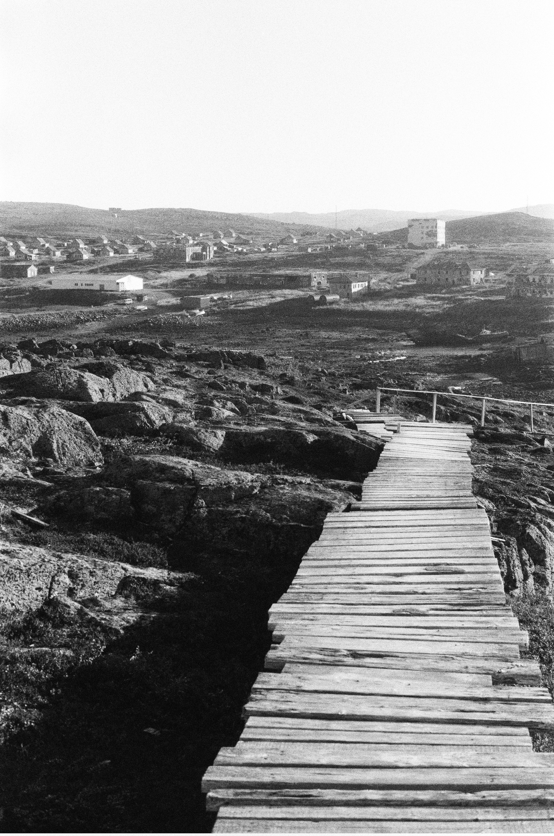 A weathered wooden walkway stretches across rocky terrain toward a distant settlement of low buildings and houses.