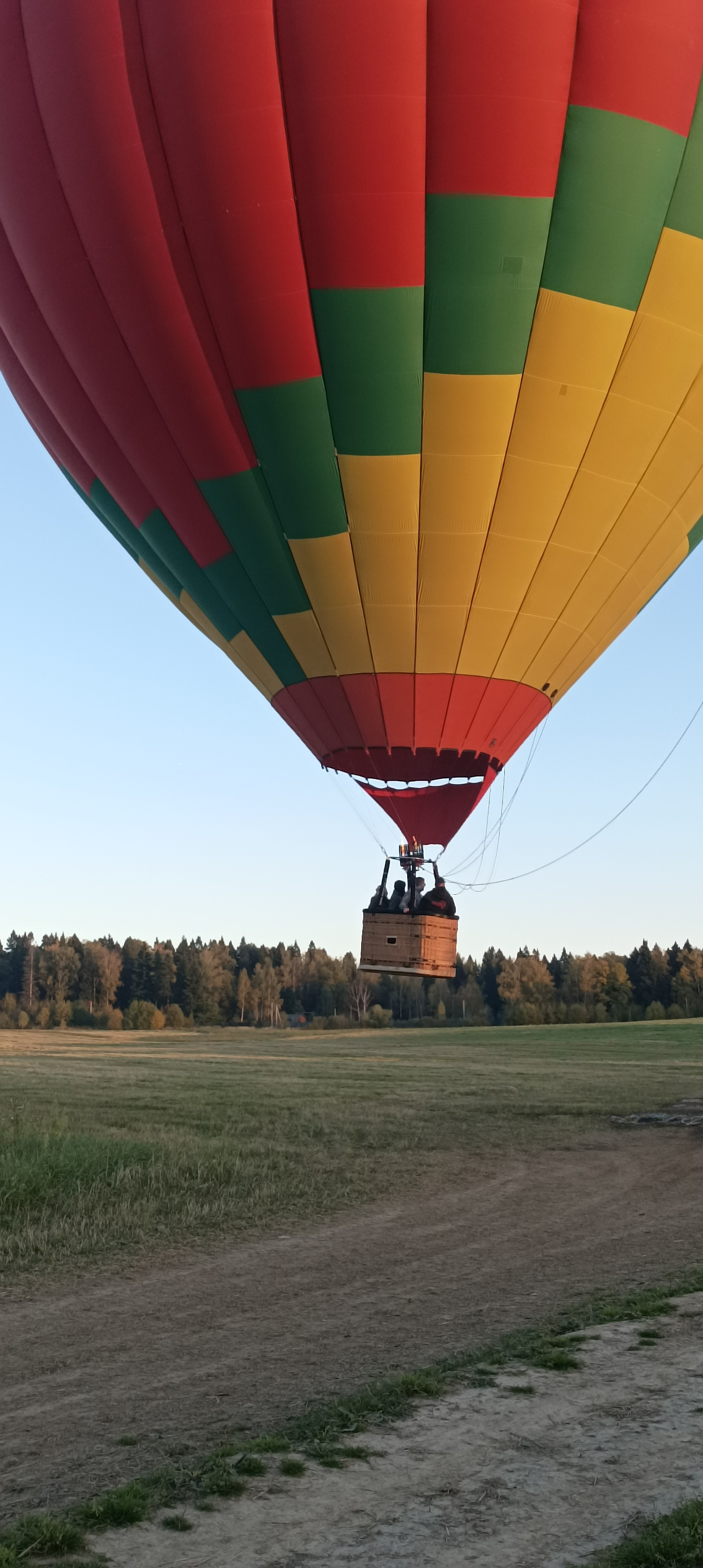 A colorful hot air balloon with red, green, and yellow patches is floating just above a grassy field near a forested area.