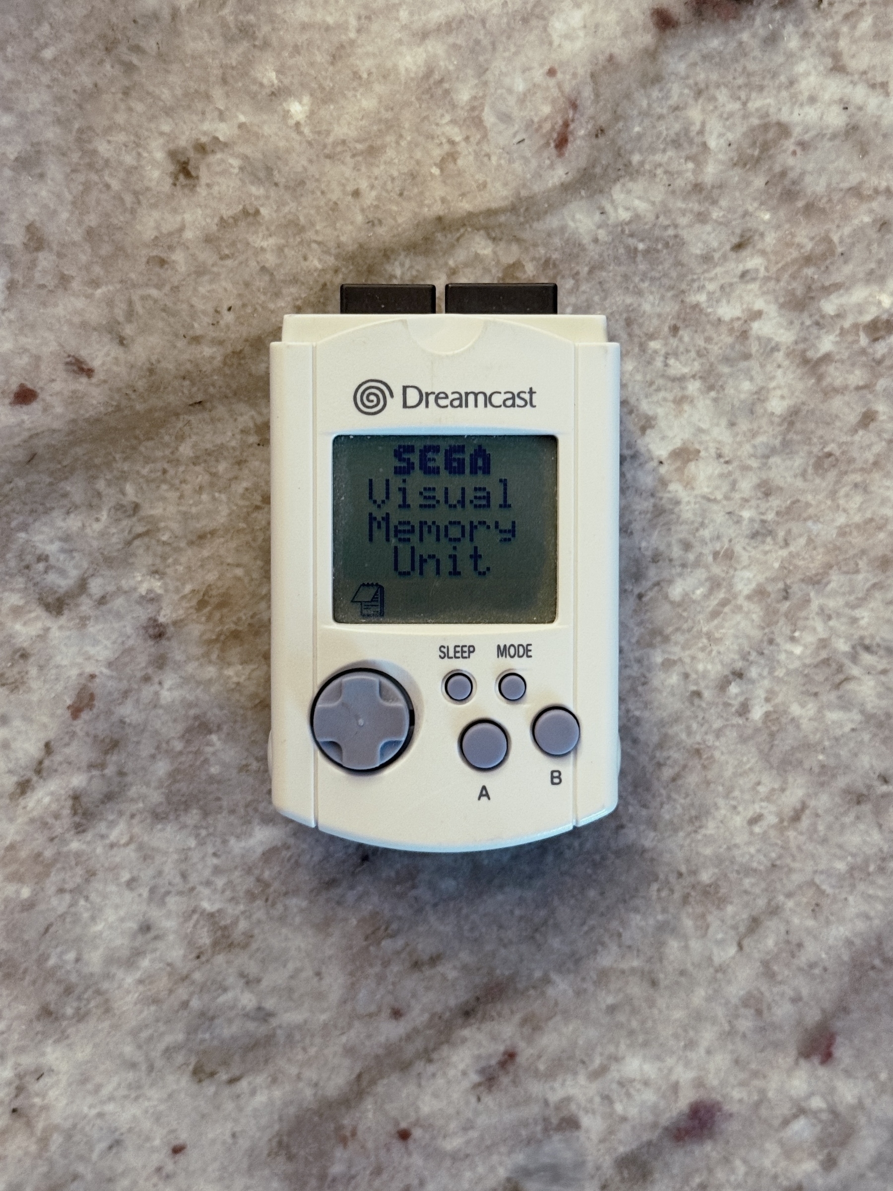 A Dreamcast Visual Memory Unit (VMU) is displayed on a textured surface.