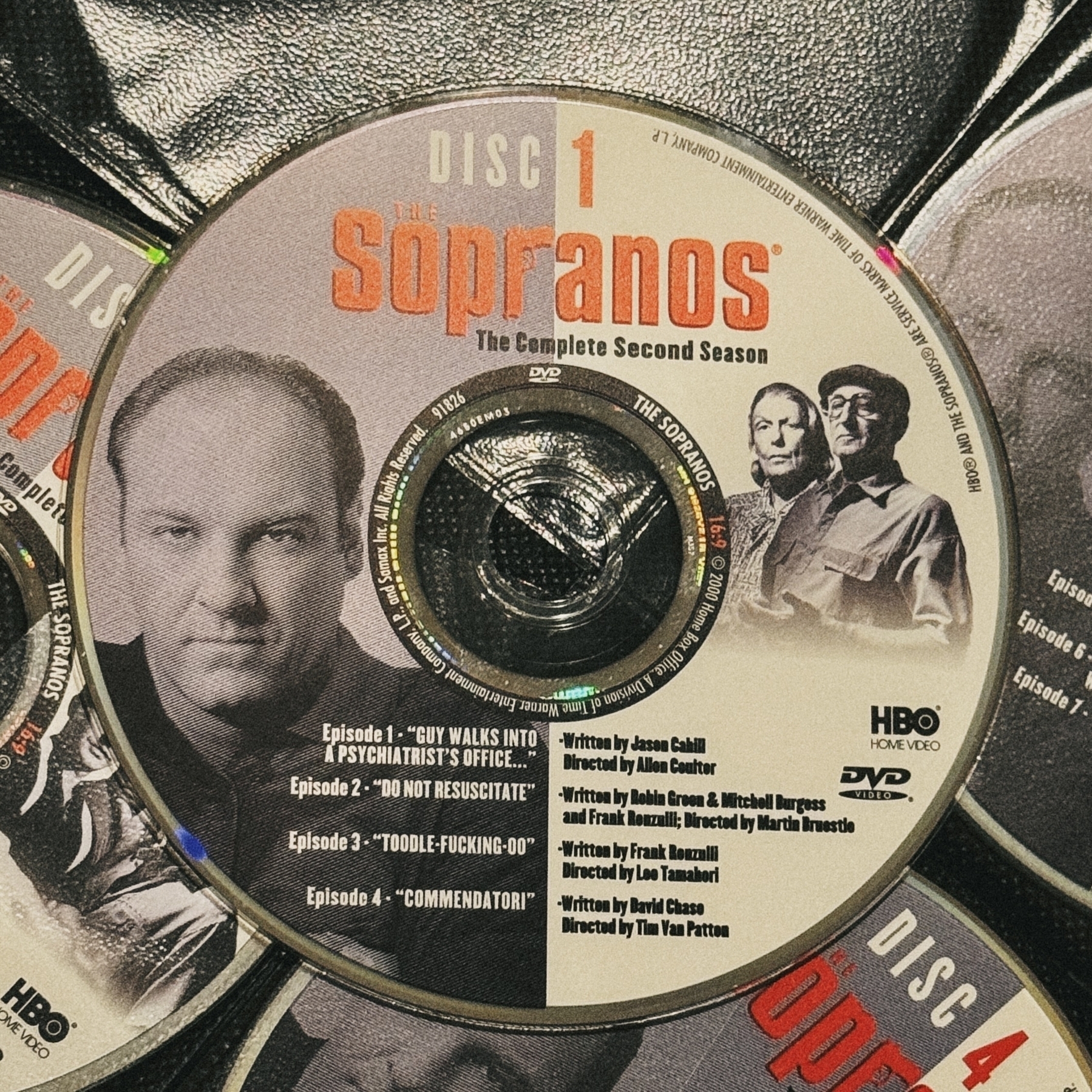 DVD disc of the first episode of the sopranos sitting atop a grid of other discs in an old black disc portfolio booklet