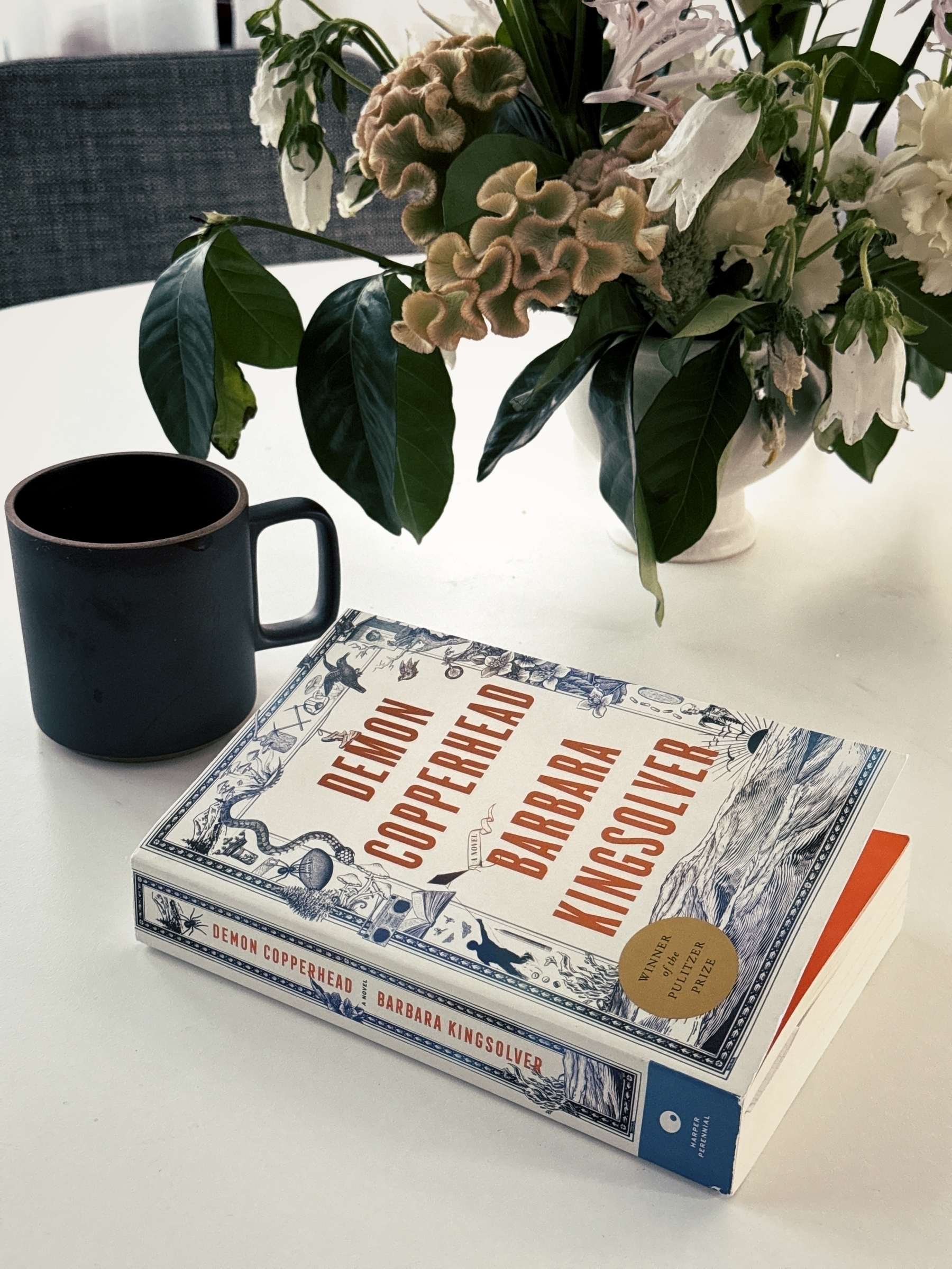 A book titled "Demon Copperhead" by Barbara Kingsolver is on a white table alongside a dark mug and a vase of flowers.