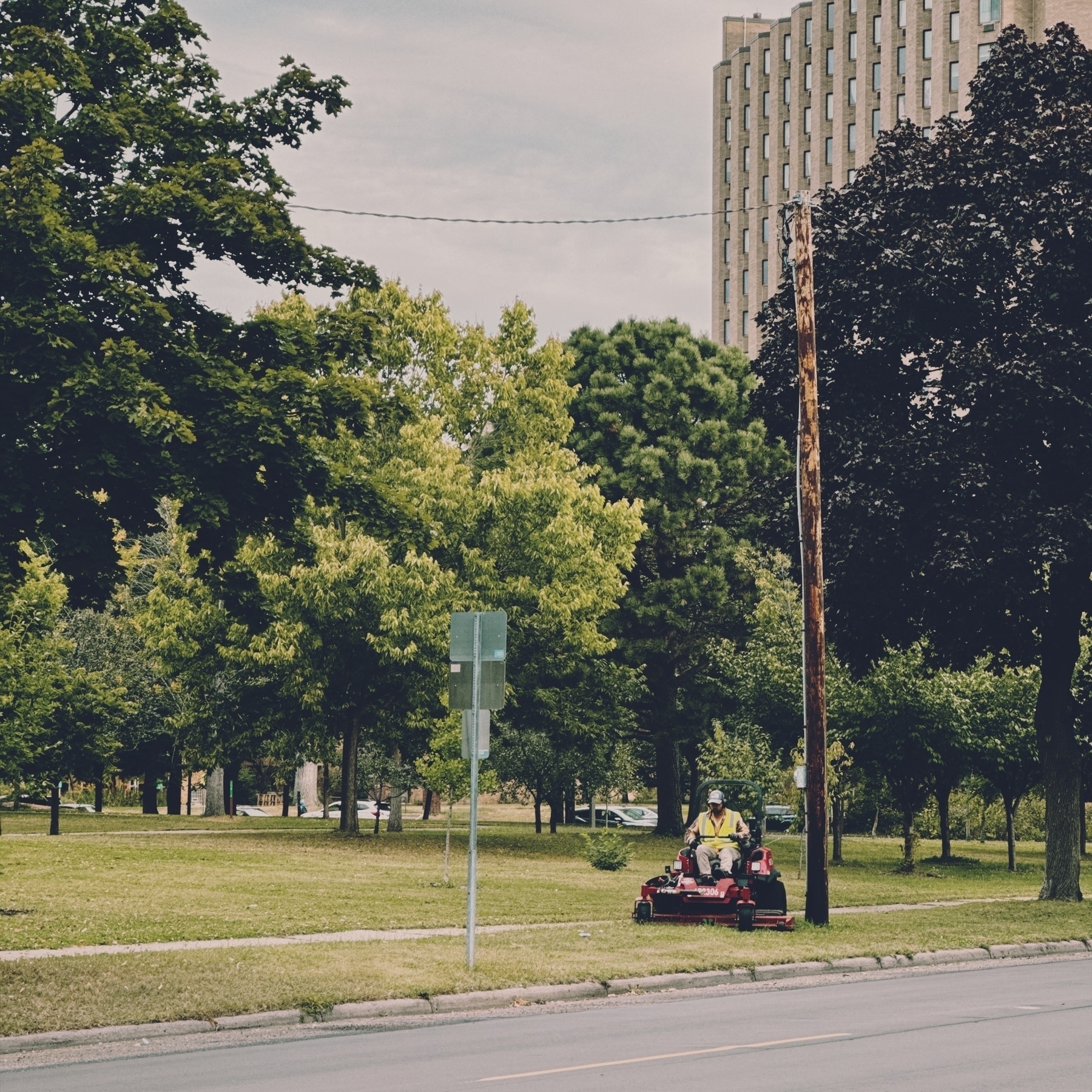 A person is operating a lawnmower on a grassy area with trees and a tall building in the background.