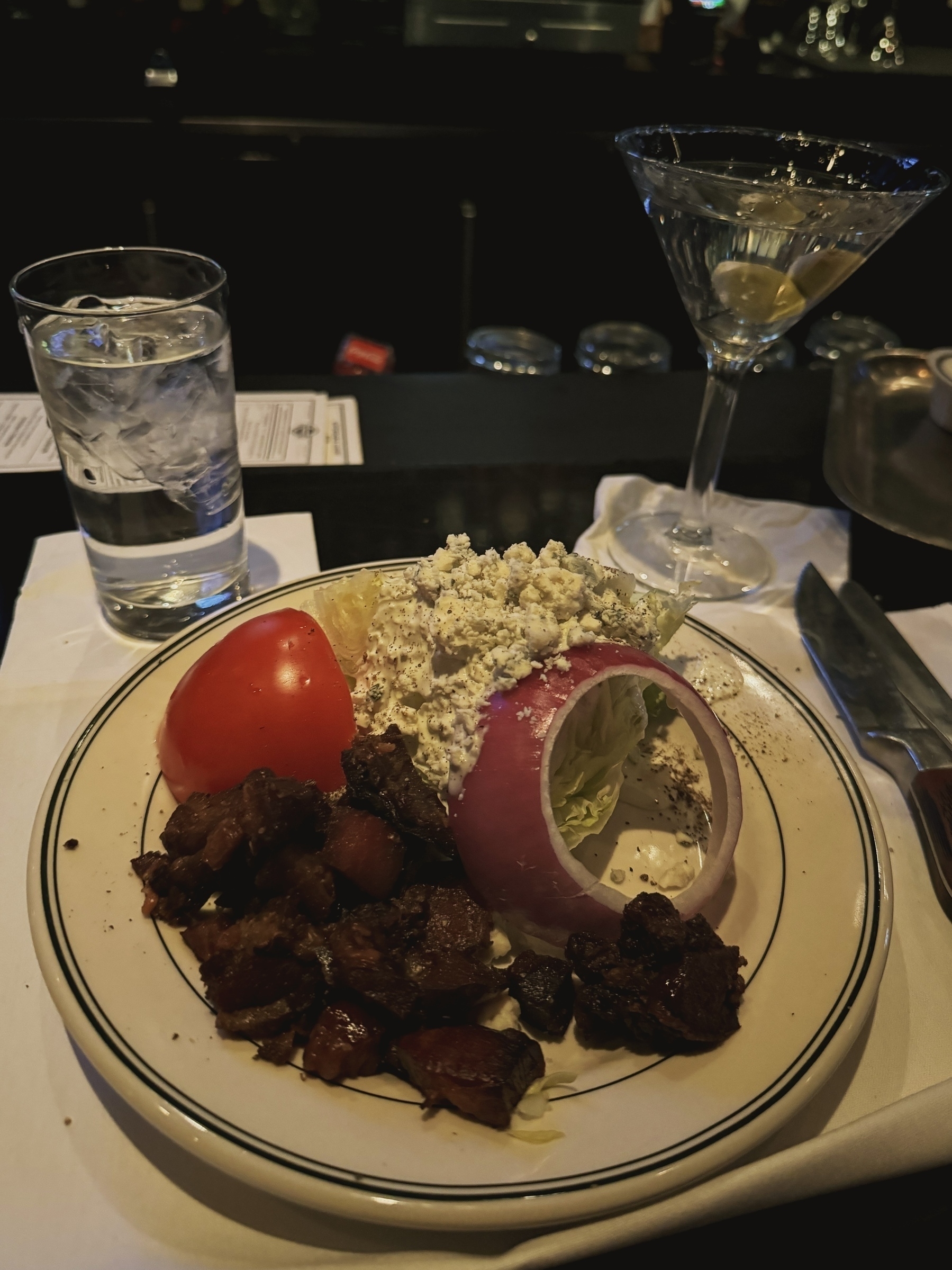 A wedge salad with an absurd amount of thick cut bacon bits next to a water glass and martini. 