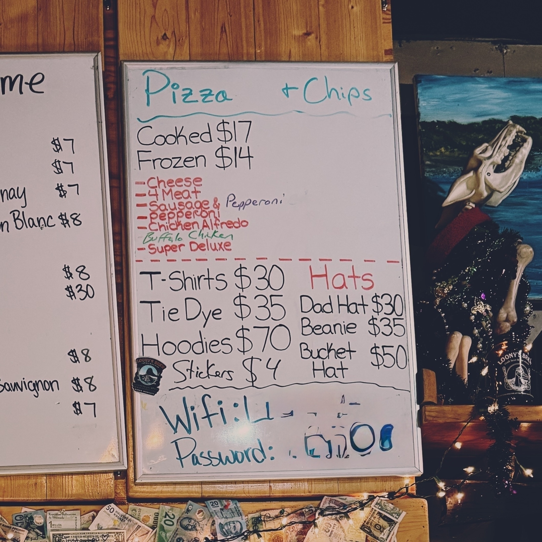 A whiteboard posted up on a wooden wall inside a bar, detailing store prices for merchandise as well as options for pizza, either cooked or frozen, with a $3 differential. 