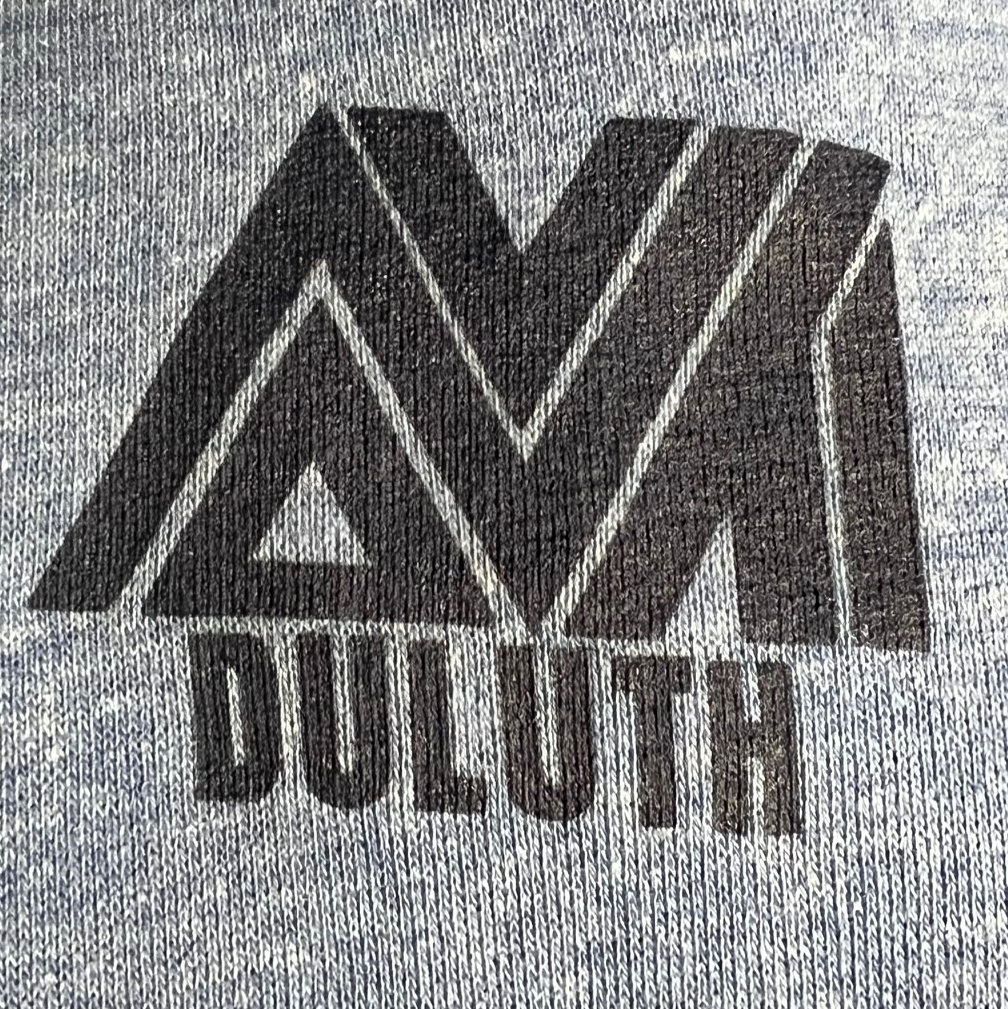 close-up photograph of a gray shirt with black logotype (jagged lines and triangles looking like a mountain) and lettering 'DULUTH'