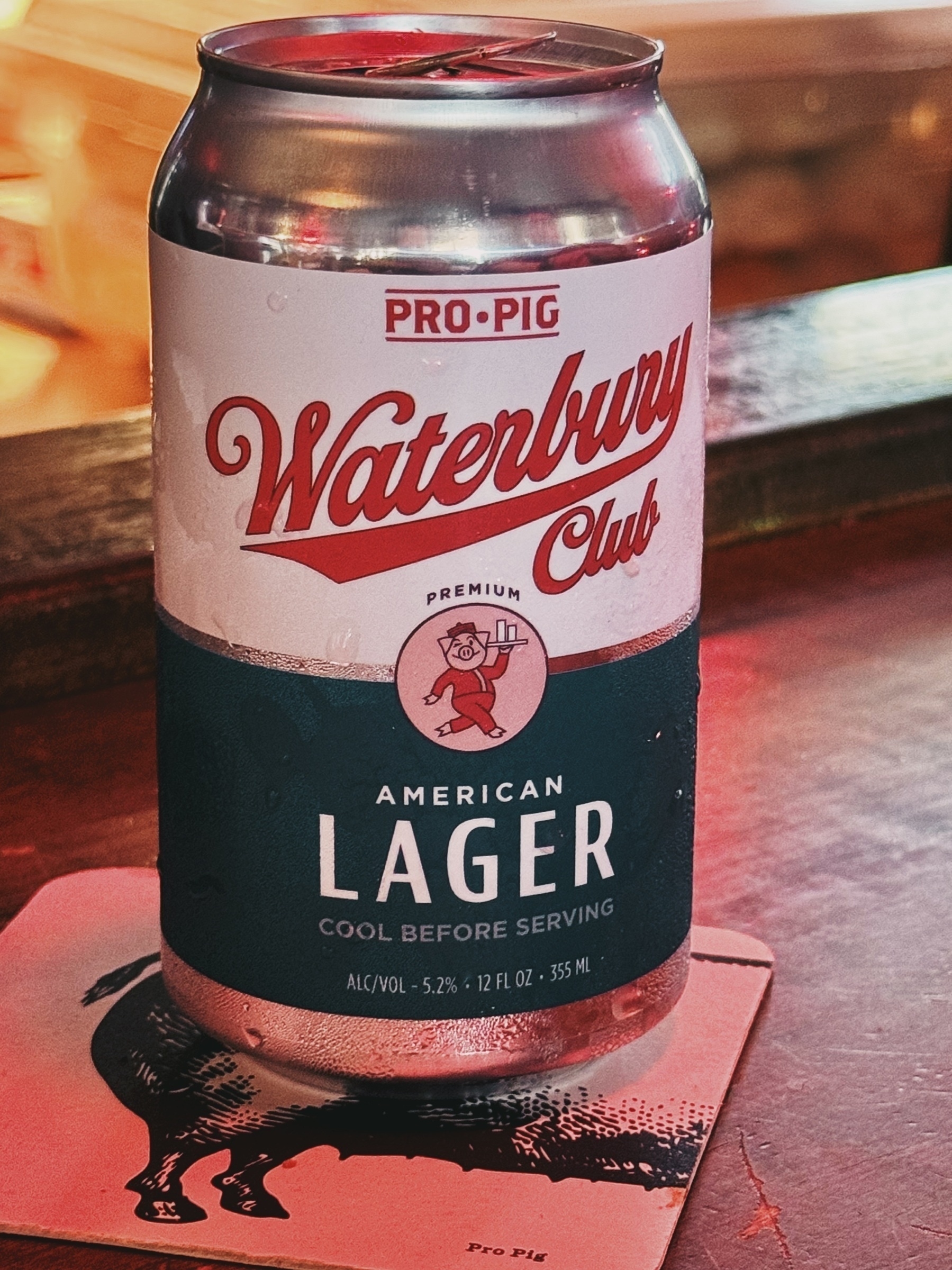 dimly lit can of beer with red cursive text called Waterbury Club Lager on a wood bar top