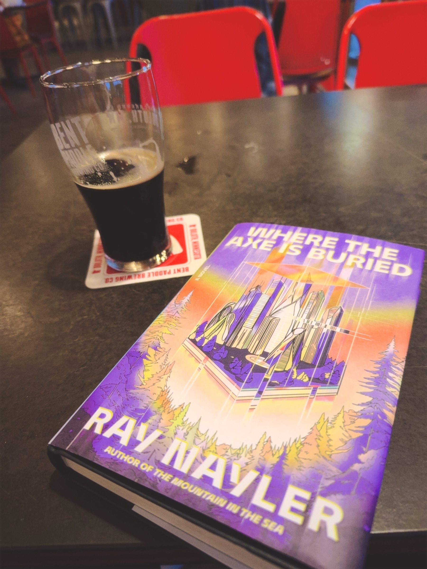 A book titled "Where the Axe is Buried" by Ray Nayler is placed on a table next to a half-full pint of dark beer.