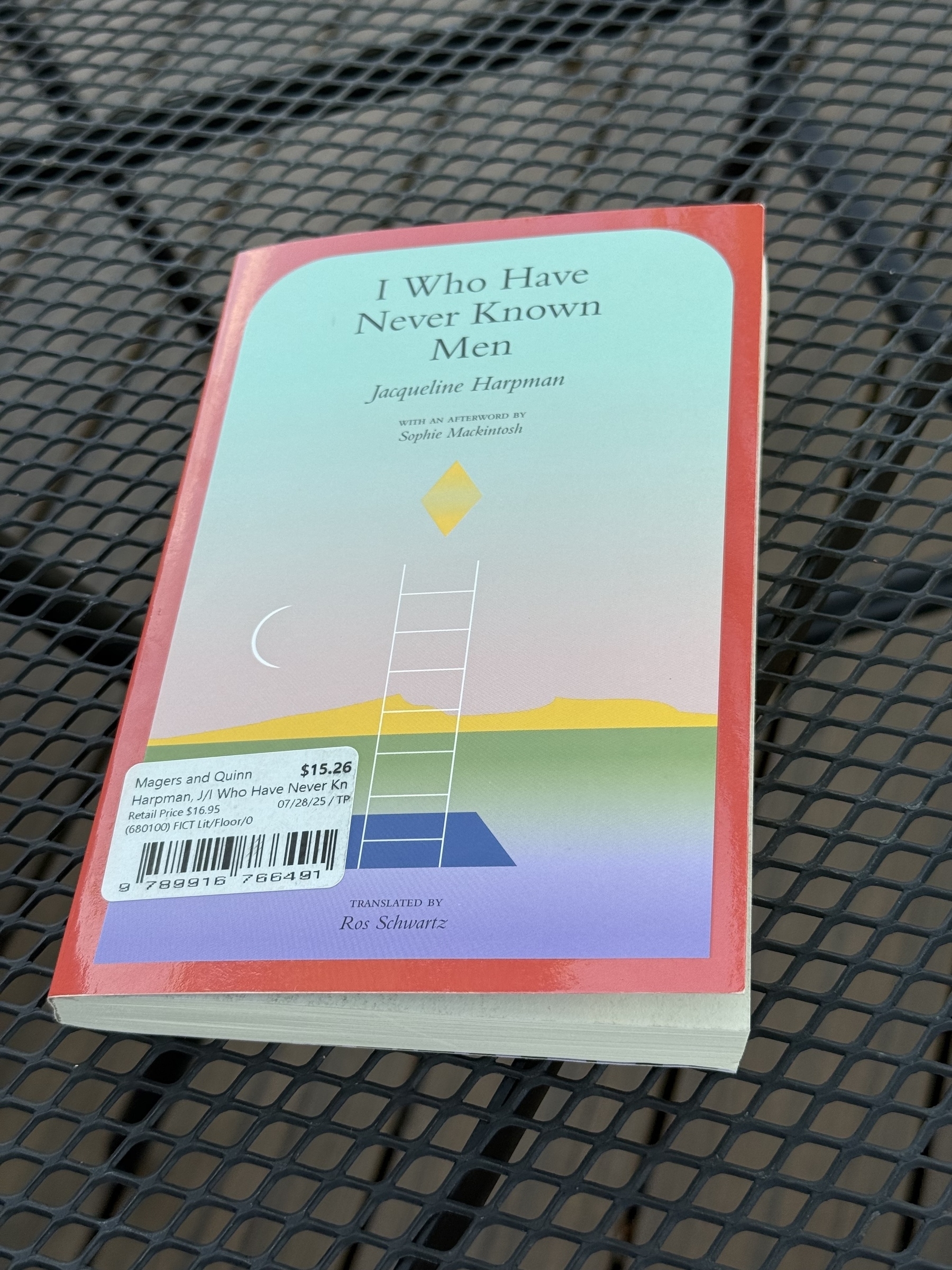A book titled "I Who Have Never Known Men" by Jacqueline Harpman is resting on a metal mesh table.