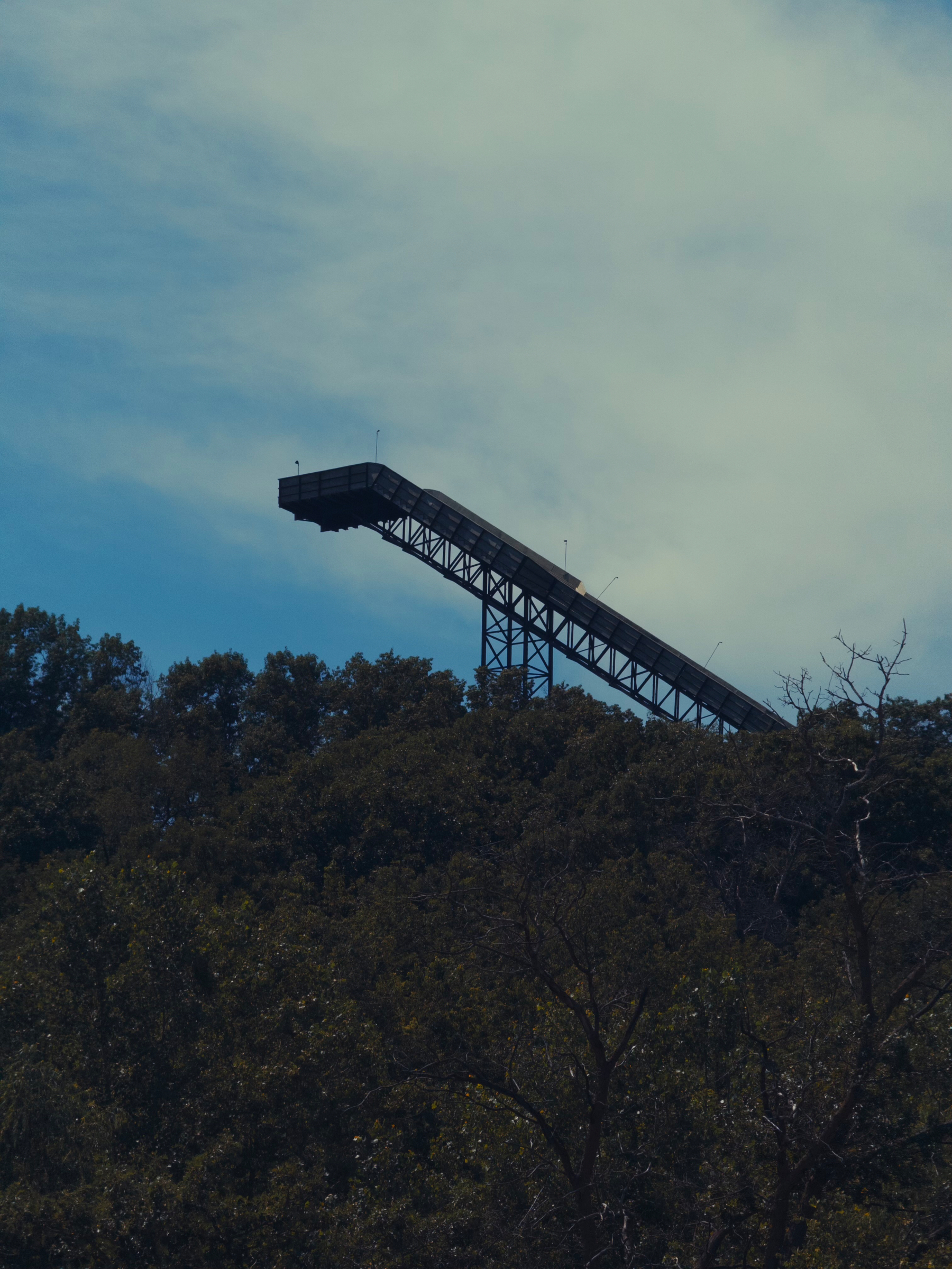 A ski jump structure extends above a wooded area under a partly cloudy sky.