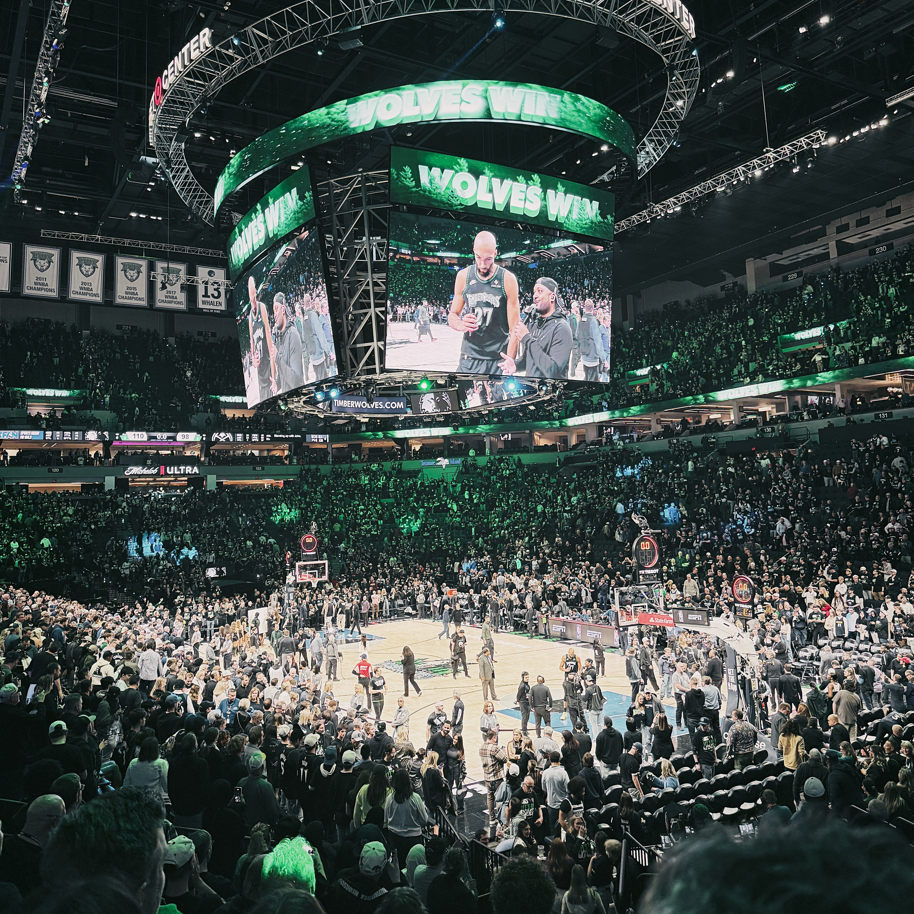 A crowded arena features a basketball game and a large screen displaying WOLVES WIN as fans watch.