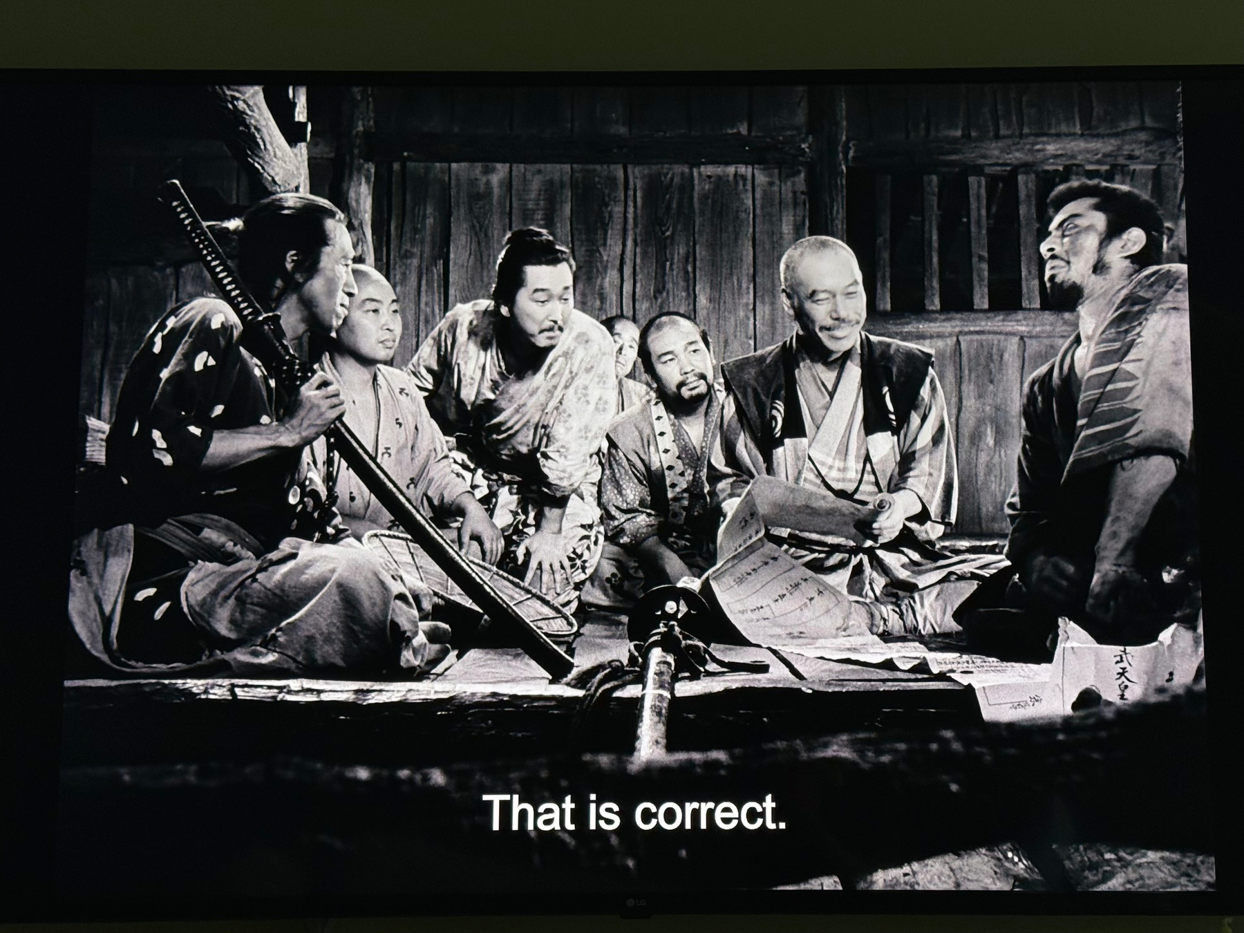 A group of people dressed in traditional samurai attire are gathered in an interior setting, focusing on a document on the floor, while one person prominently displays a sword; subtitles read That is correct. From Seven Samurai. 