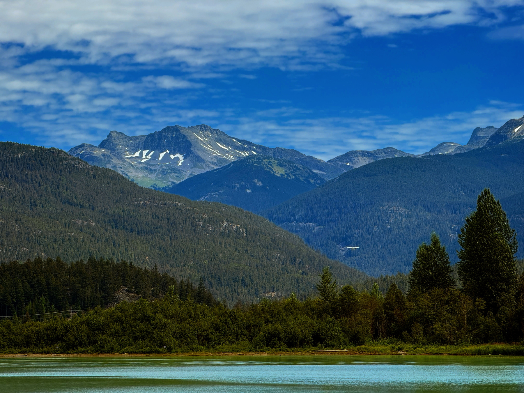 A tranquil lake is surrounded by dense green forests and towering mountains under a vibrant blue sky with scattered clouds.