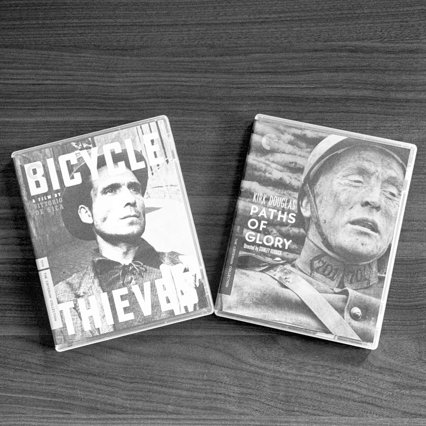 Two classic film blu-rays, Bicycle Thieves and Paths of Glory, are placed on a wooden surface. Black and White (filtered via the AgBr app)