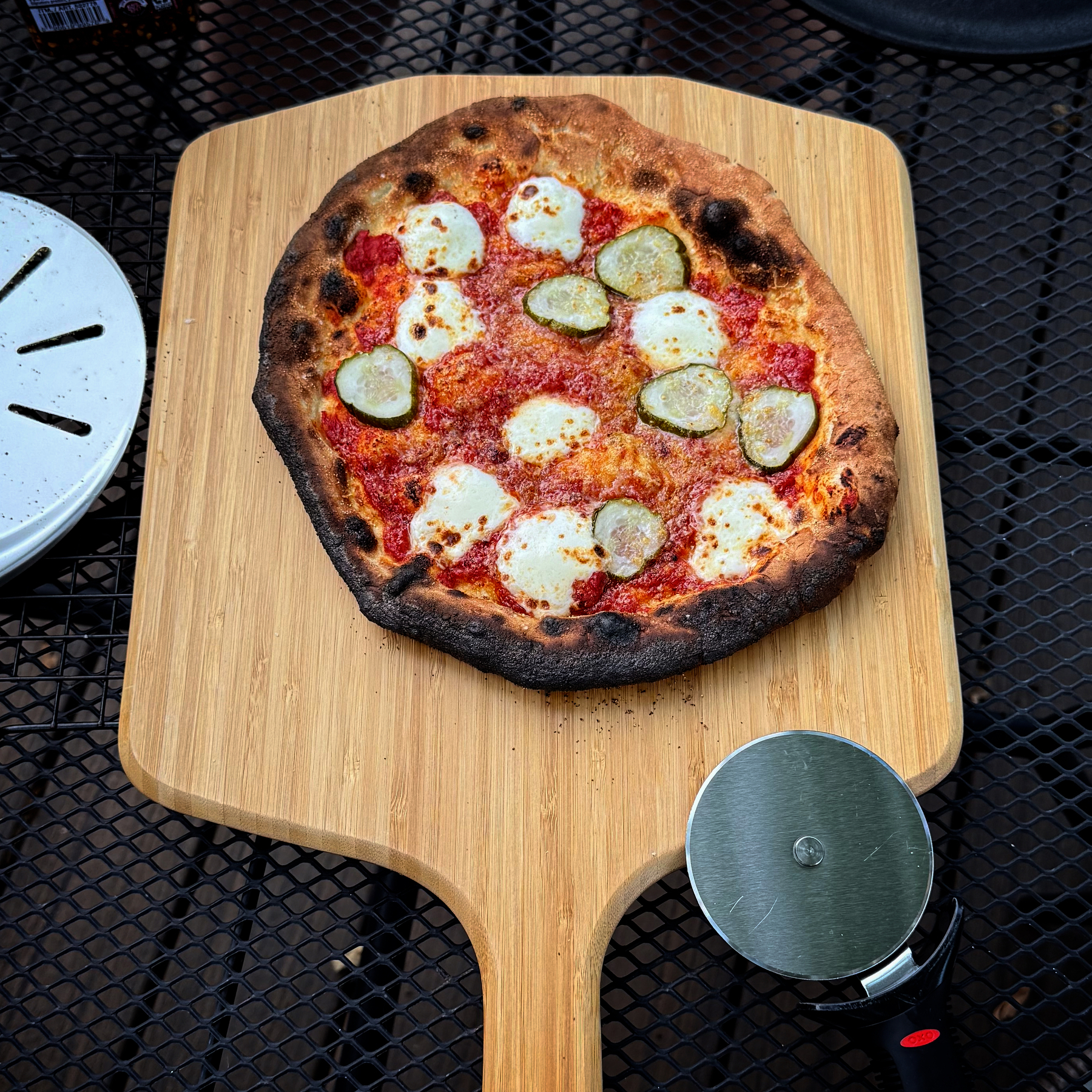 A pizza with melted cheese and slices of pickles on a wooden pizza peel, placed on a black metal mesh table along with a pizza cutter.