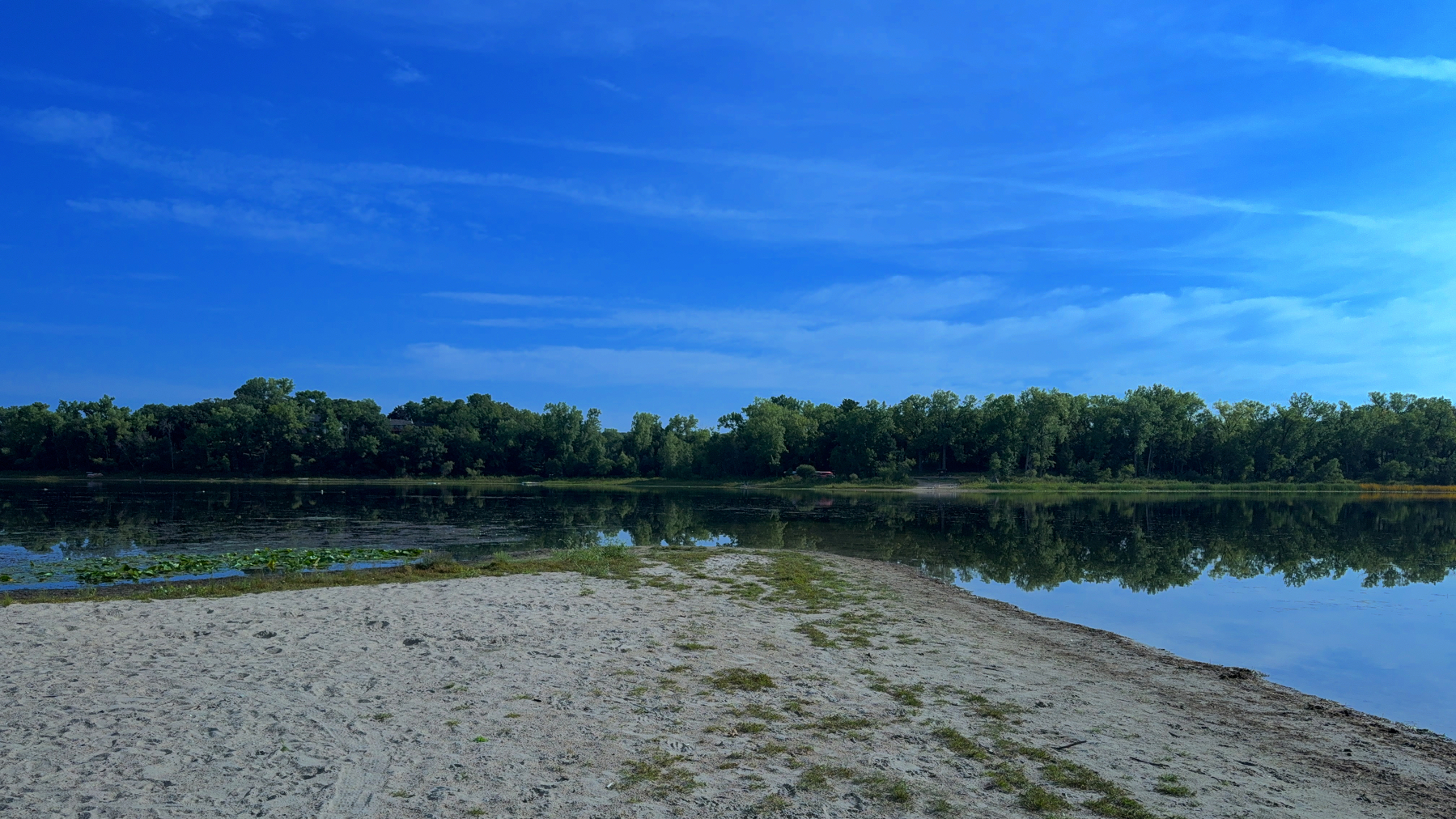A serene lake scene features a sandy shore in the foreground, calm water reflecting the surrounding trees, and a partly cloudy blue sky above.