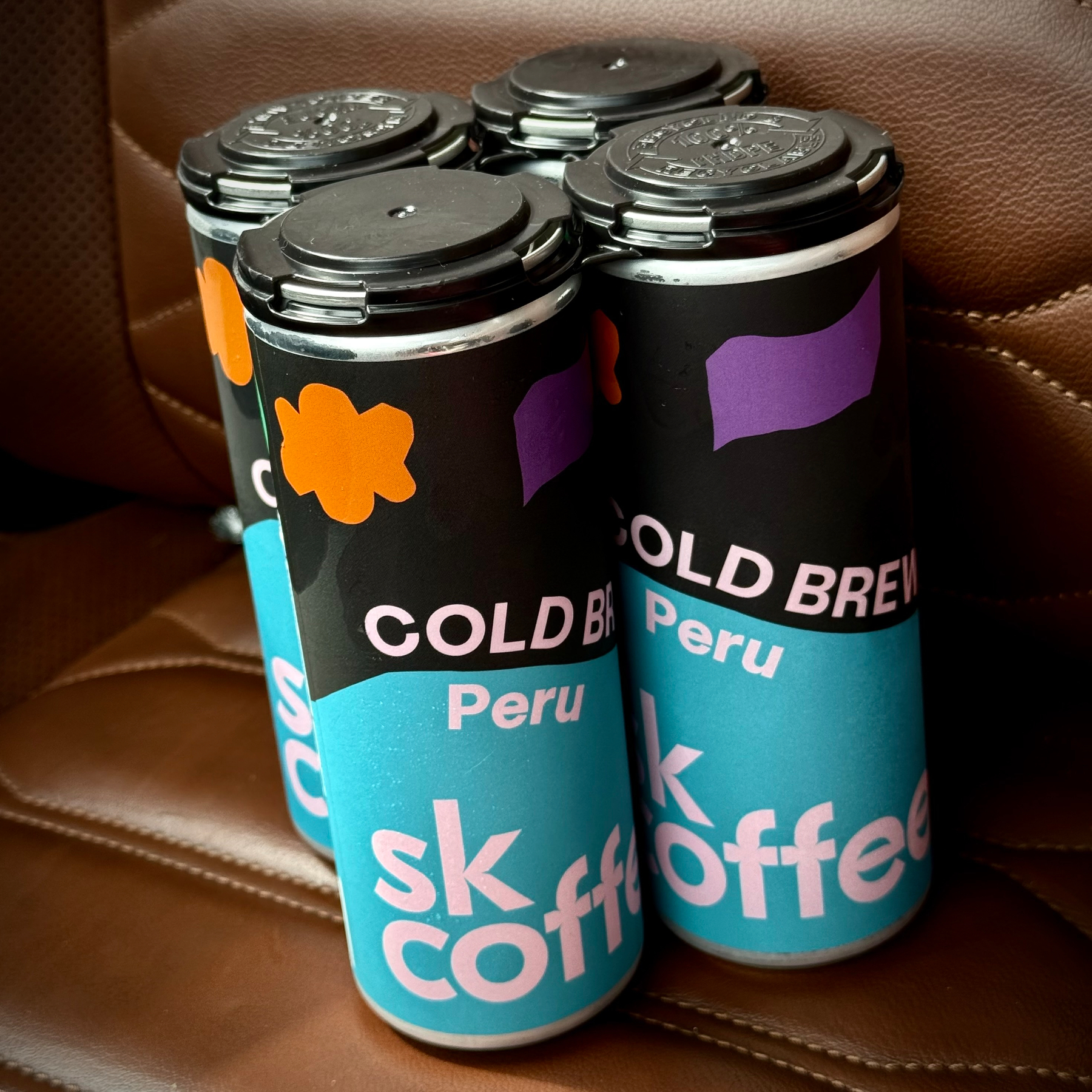 A four-pack of tall cans labeled Cold Brew Peru, by SK Coffee is placed on a brown leather surface.