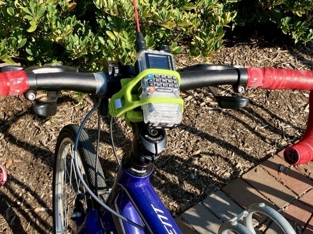 Blue bike at a bike rack with a clear handheld transceiver mounted to the handle bars. The handle bars are wrapped in red tape.