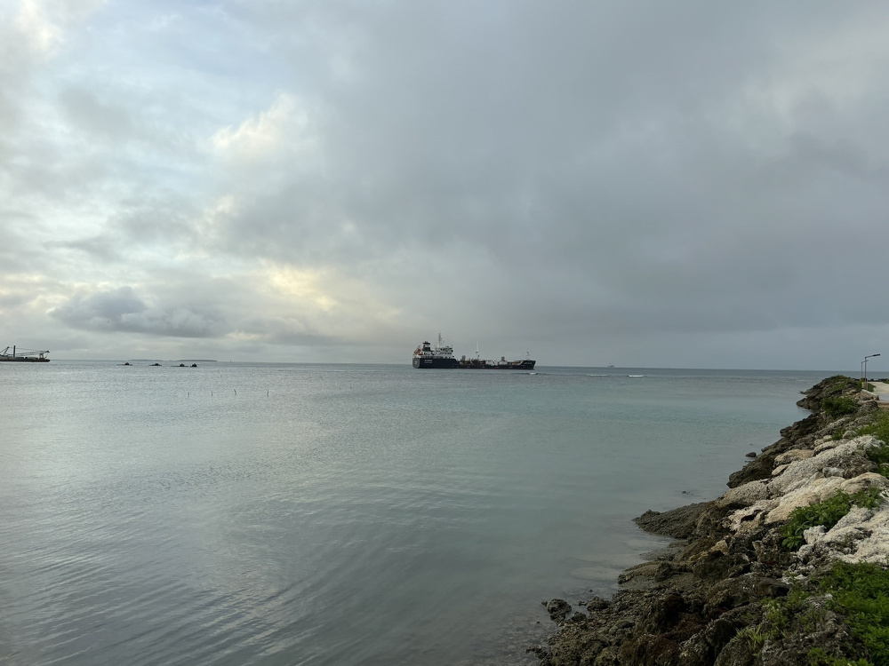 A large ship is sailing on a calm sea under a cloudy sky, with a rocky shoreline visible in the foreground.