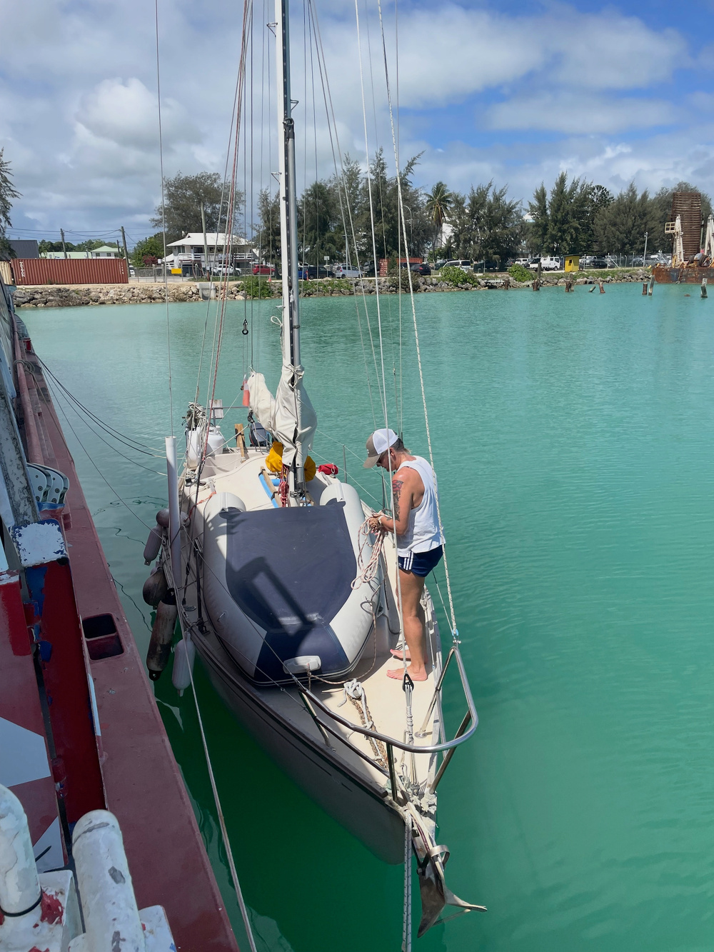 A person stands on a docked sailboat with calm turquoise water and trees in the background.