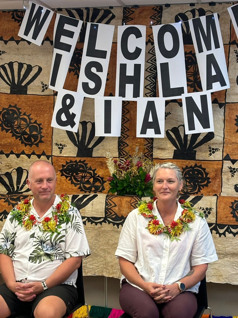 Two people are sitting in front of a decorative backdrop with a floral garland around their necks, beneath a Welcome Ishla & Ian sign.