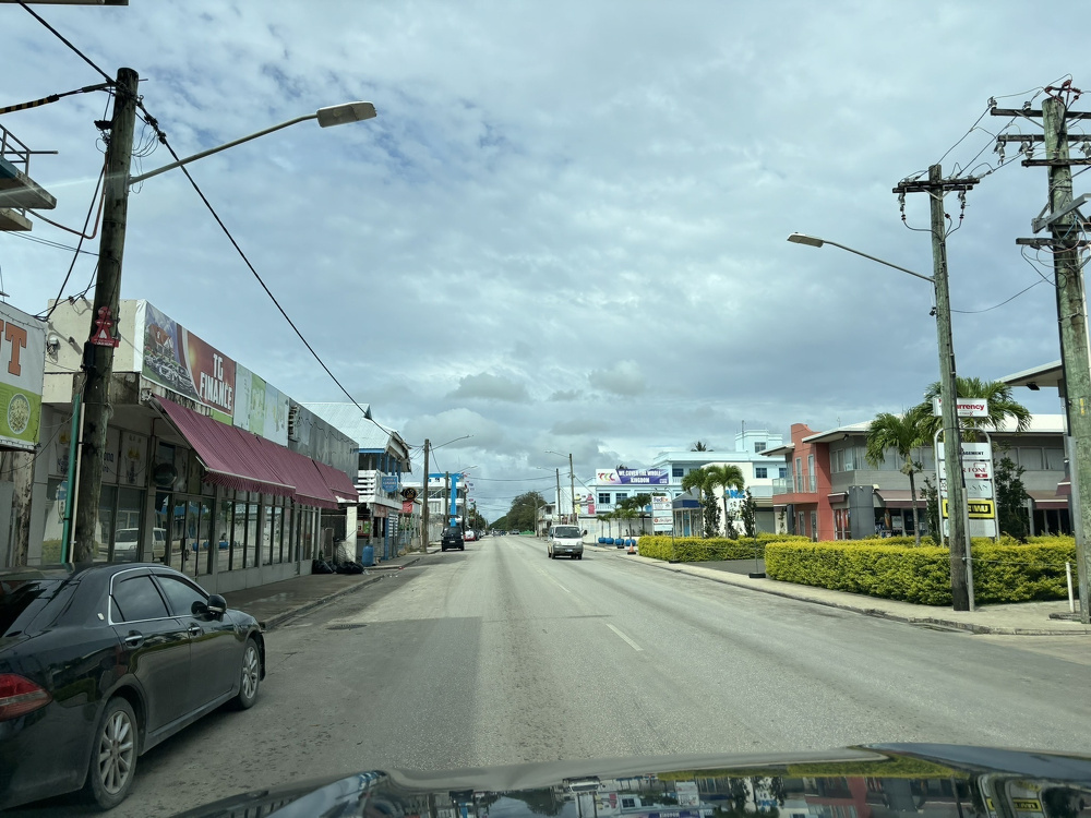 A two-lane road is flanked by commercial buildings, cars, and utility poles under a cloudy sky.