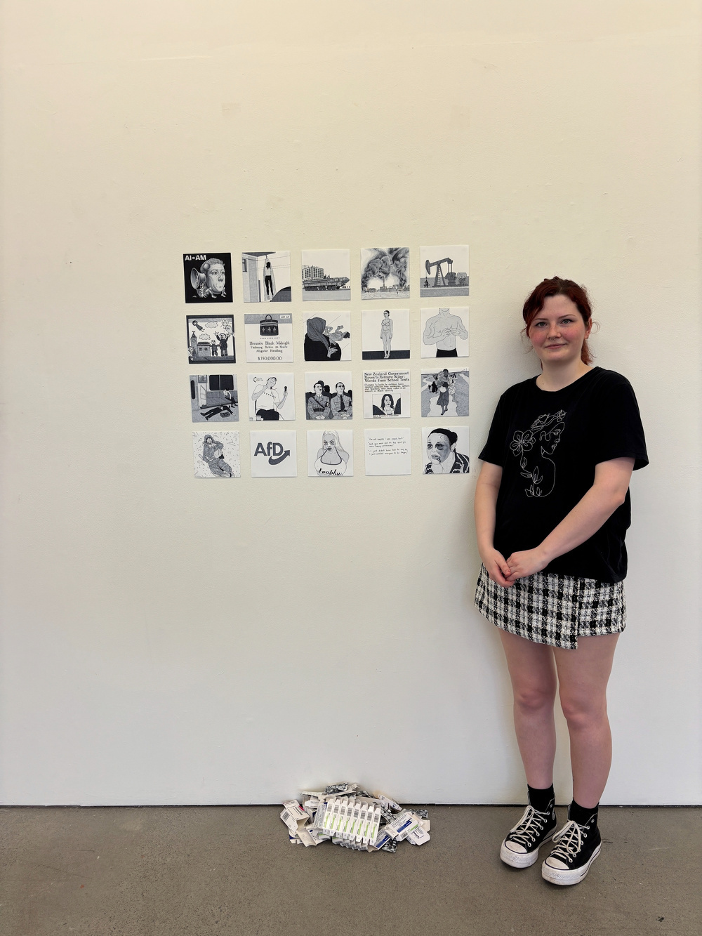 A person is standing beside a wall displaying a grid of artistic images, with a pile of materials on the floor below.