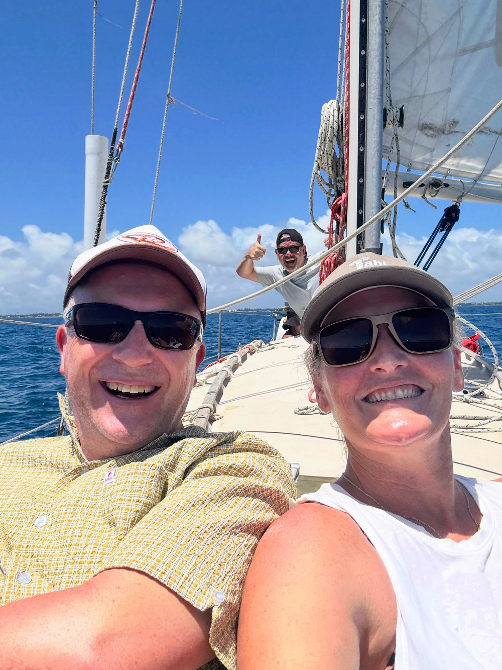 A smiling couple is taking a selfie on a sailing boat with a man in the background giving a thumbs-up.