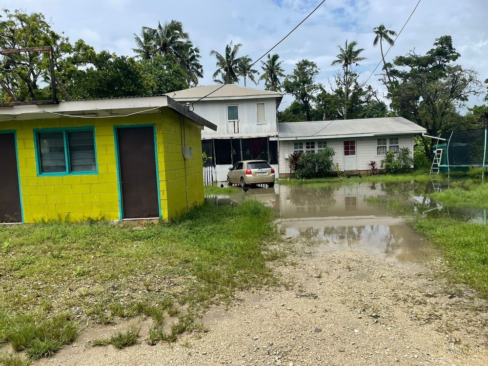 A small, waterlogged area with two houses, a car, and a trampoline, surrounded by grass and palm trees.