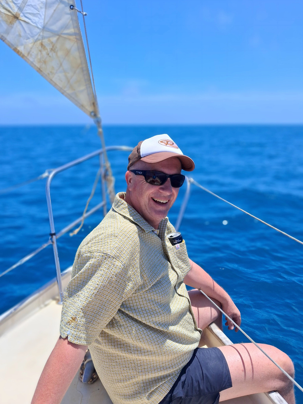 A person wearing sunglasses and a cap is smiling while sitting on a sailboat in the ocean.