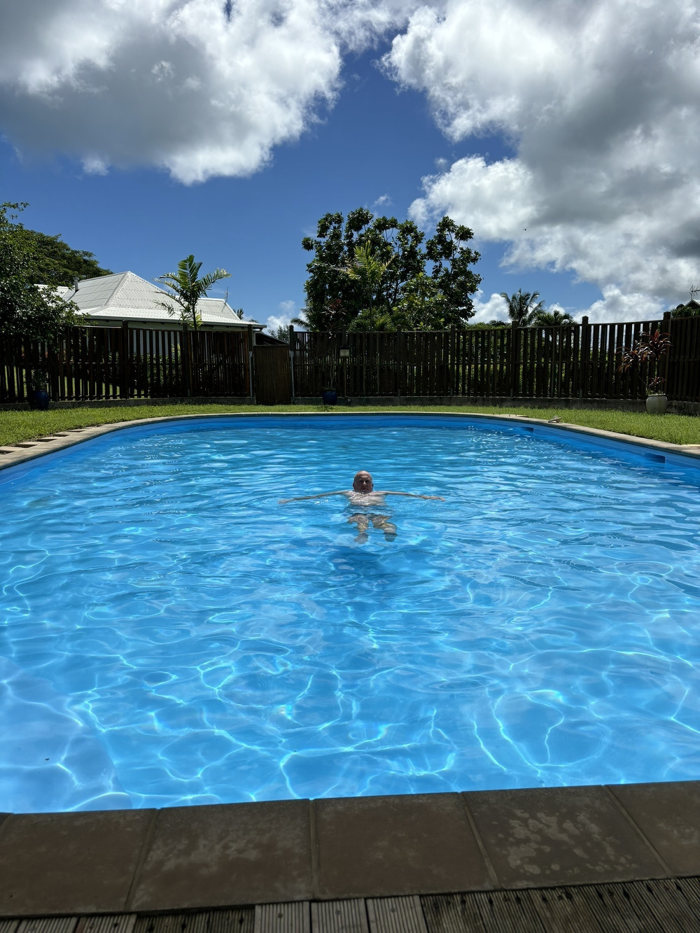 A person wearing a red and white cap is swimming in a bright blue pool under a partly cloudy sky, with trees and a fence surrounding the area.