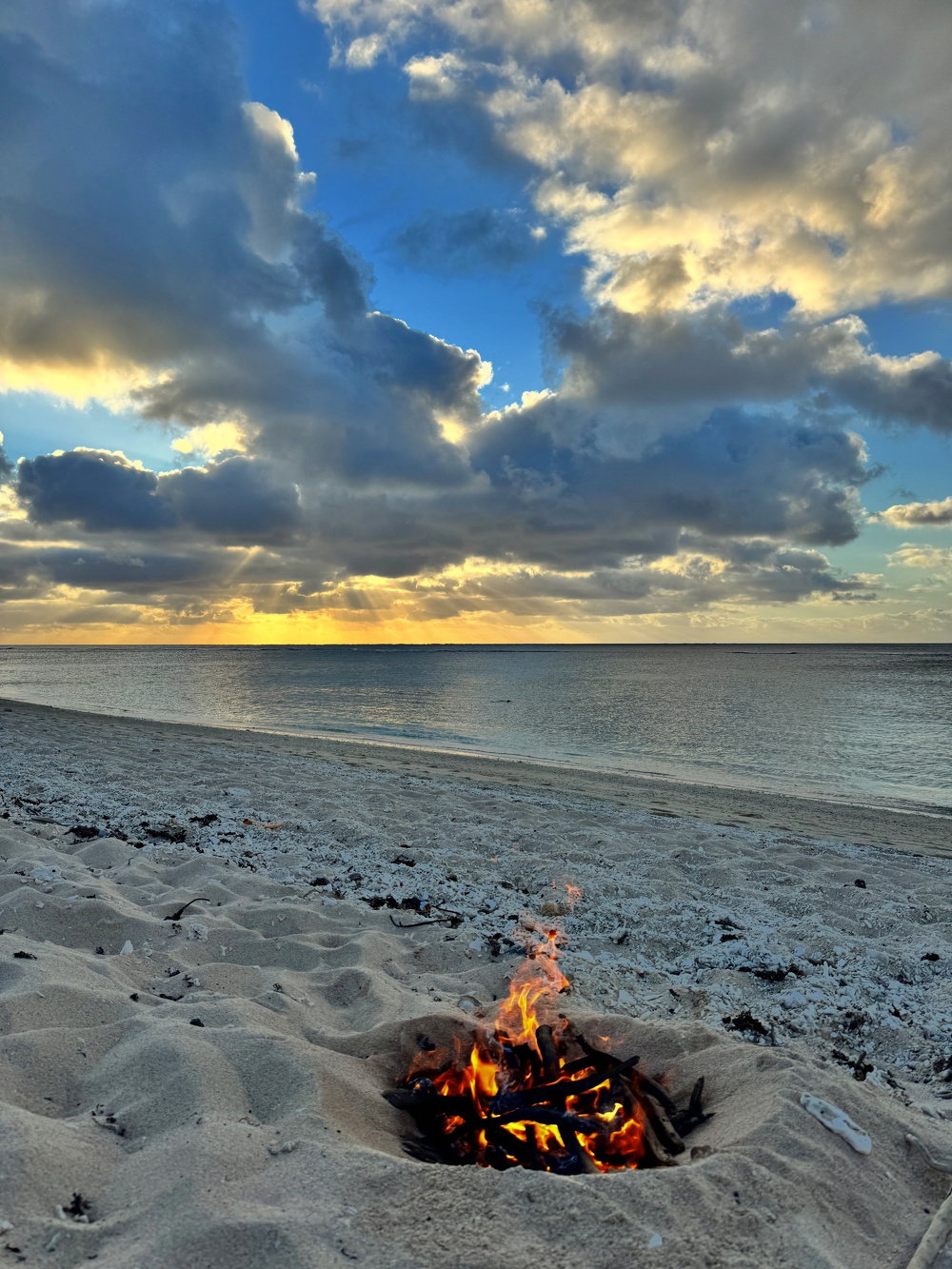A small fire burns in a sandy pit on a beach at sunset, with dramatic clouds and calm ocean in the background.