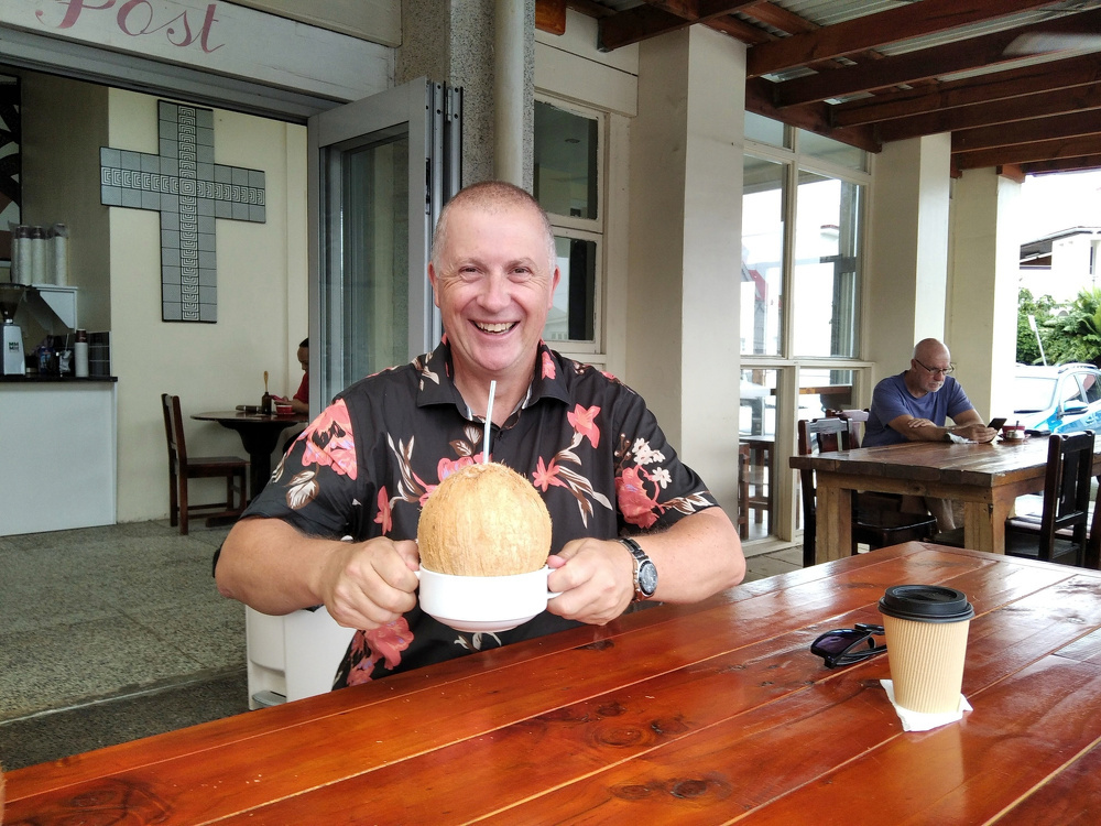 A person is smiling while holding a coconut drink with a straw at an outdoor table.