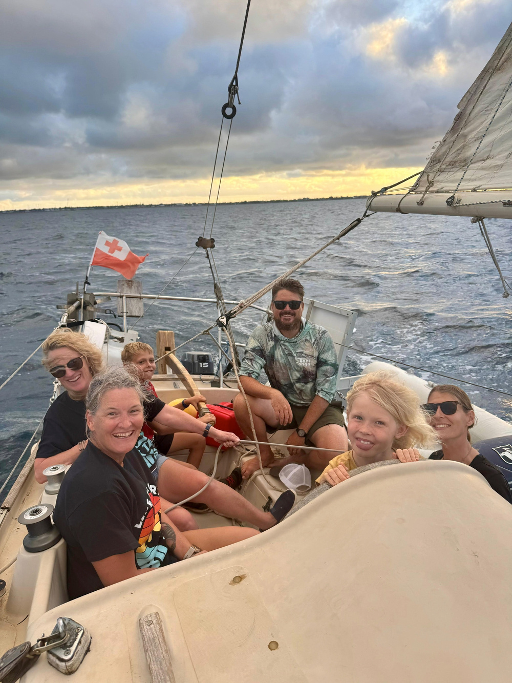A group of six people are smiling and sailing on a boat with a cloudy sky and ocean in the background.
